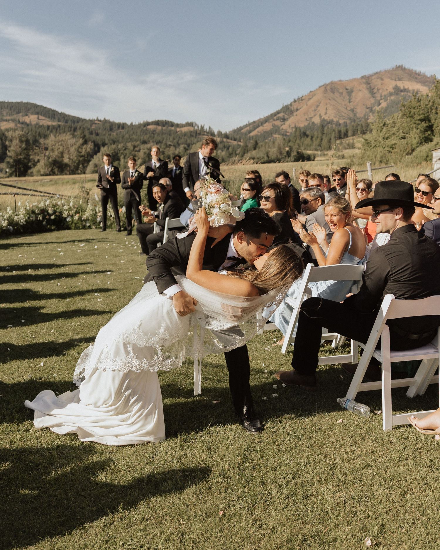 Groom dips bride for a romantic kiss at outdoor mountain wedding ceremony while guests cheer.
