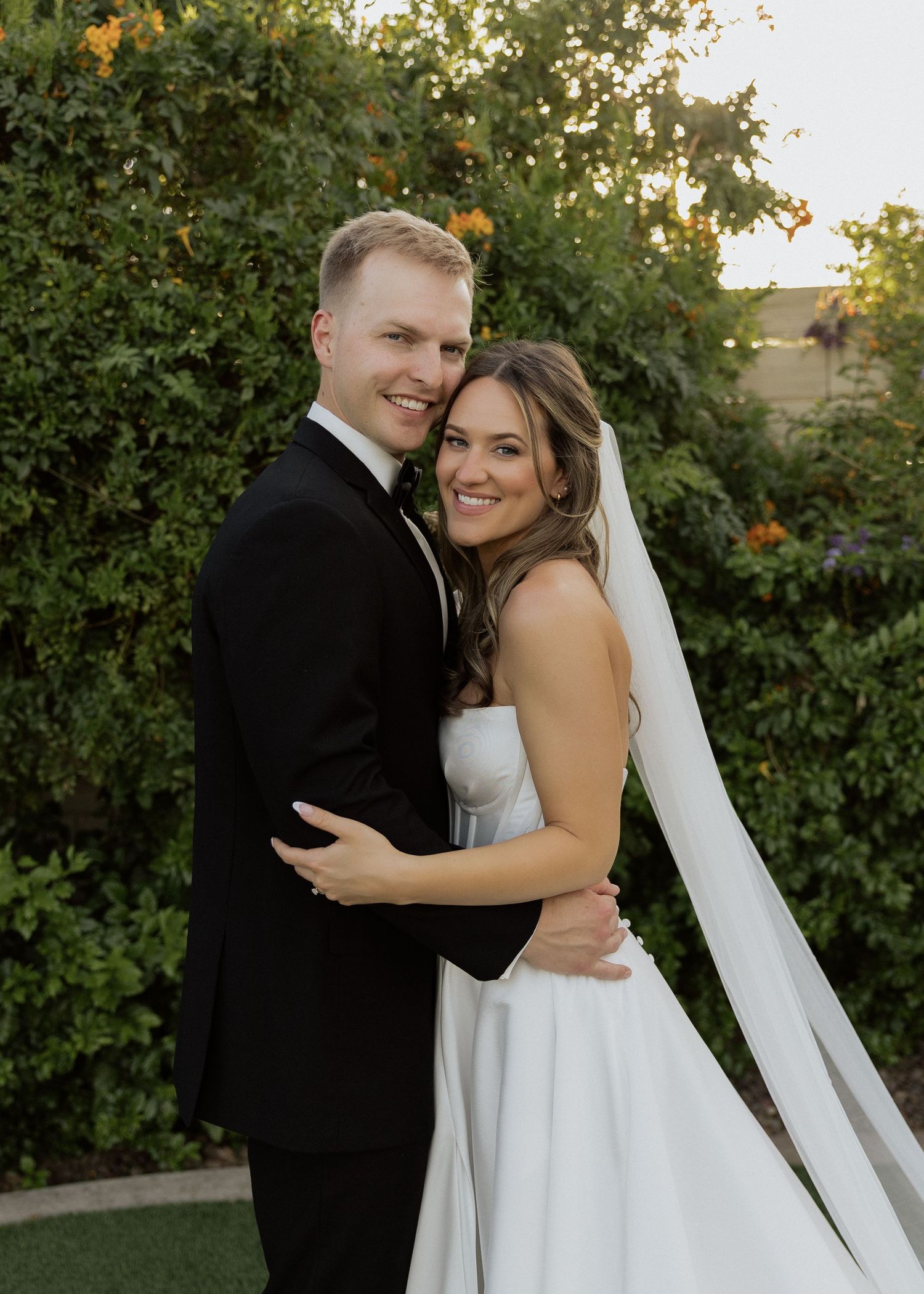 A smiling bride and groom embrace outdoors in wedding attire, with lush green foliage in the background.
