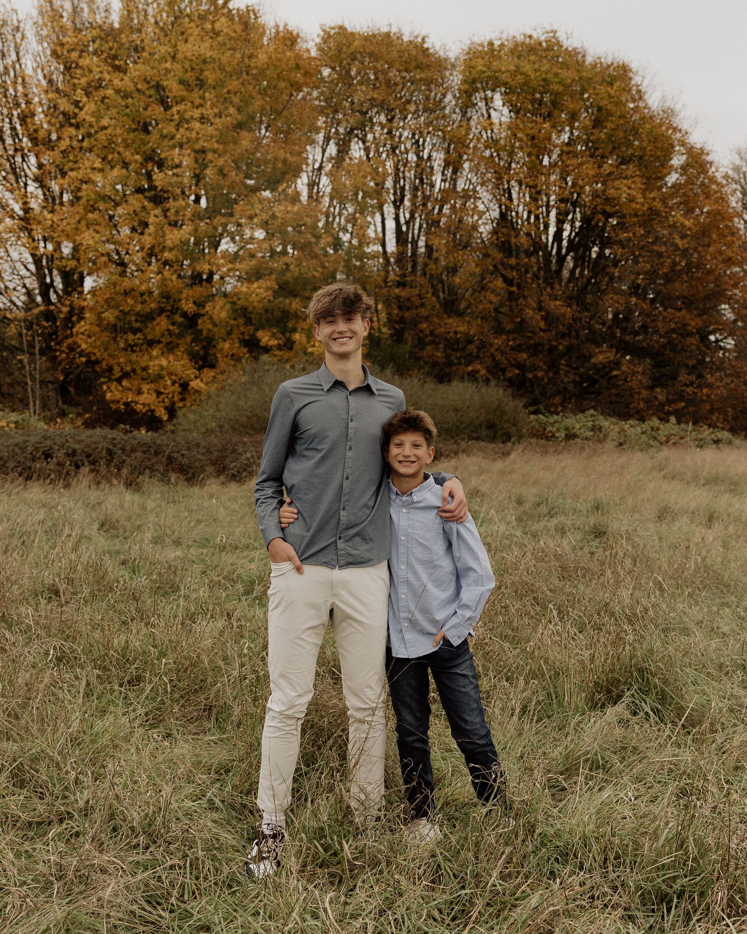 Two people standing together in a field during autumn with golden trees behind them.