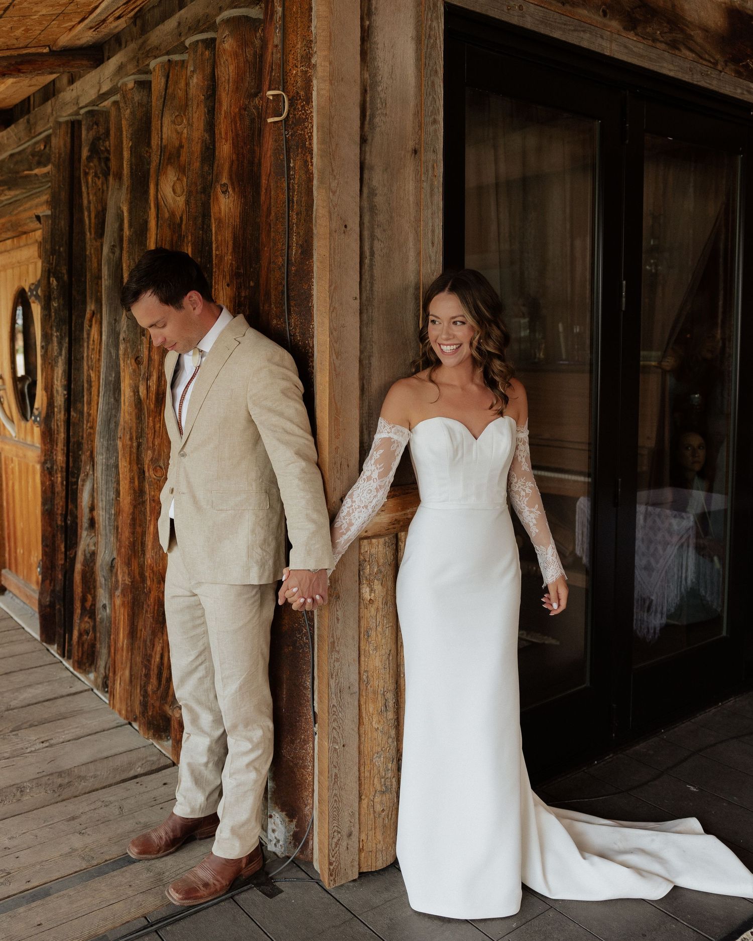 Romantic first look moment of couple holding hands against rustic wooden building exterior.