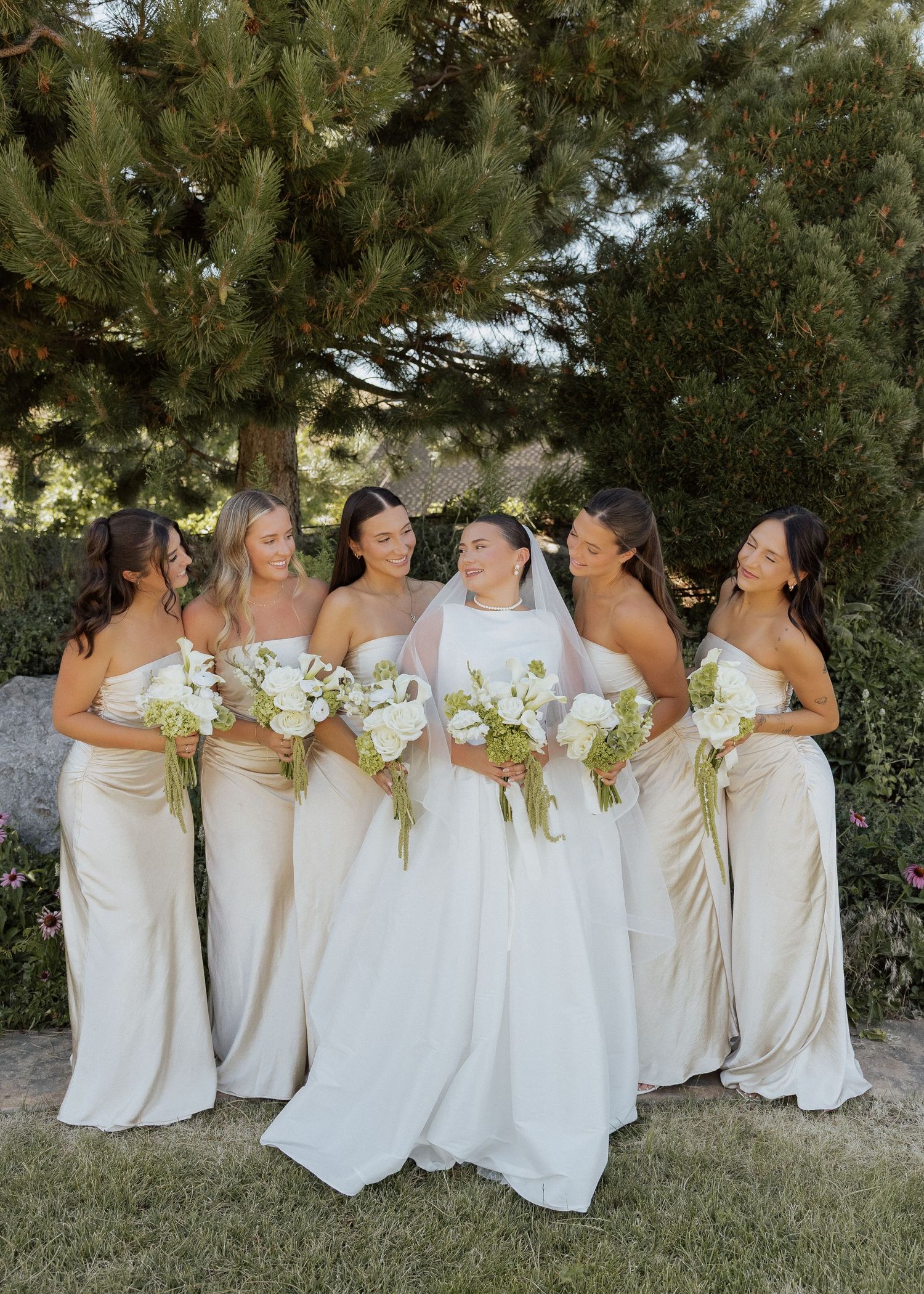 Bride in white gown and veil stands with four bridesmaids in champagne satin dresses holding white floral bouquets outdoors.