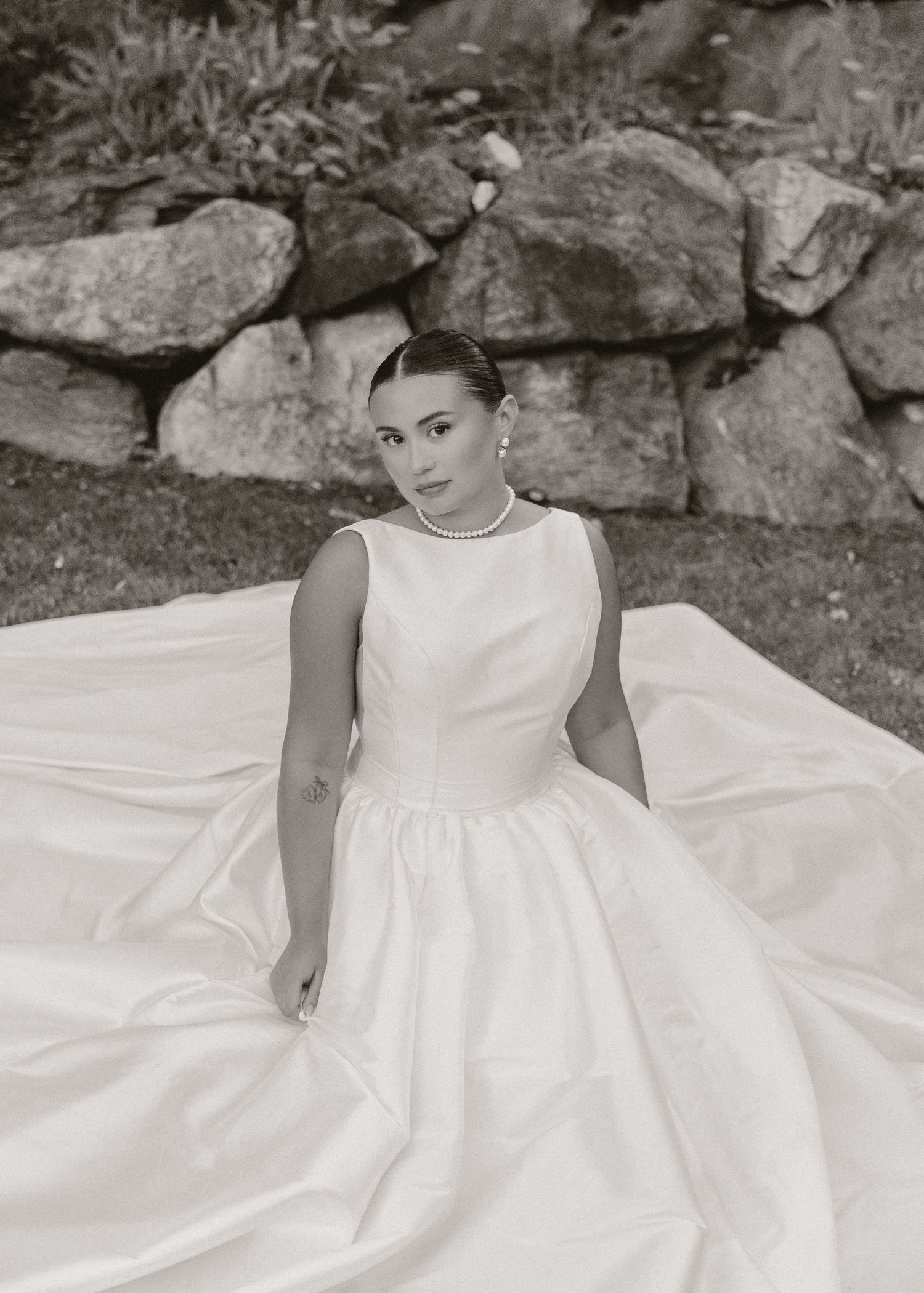 Black and white photo of a bride in a elegant ball gown wedding dress with pearl necklace seated outdoors.