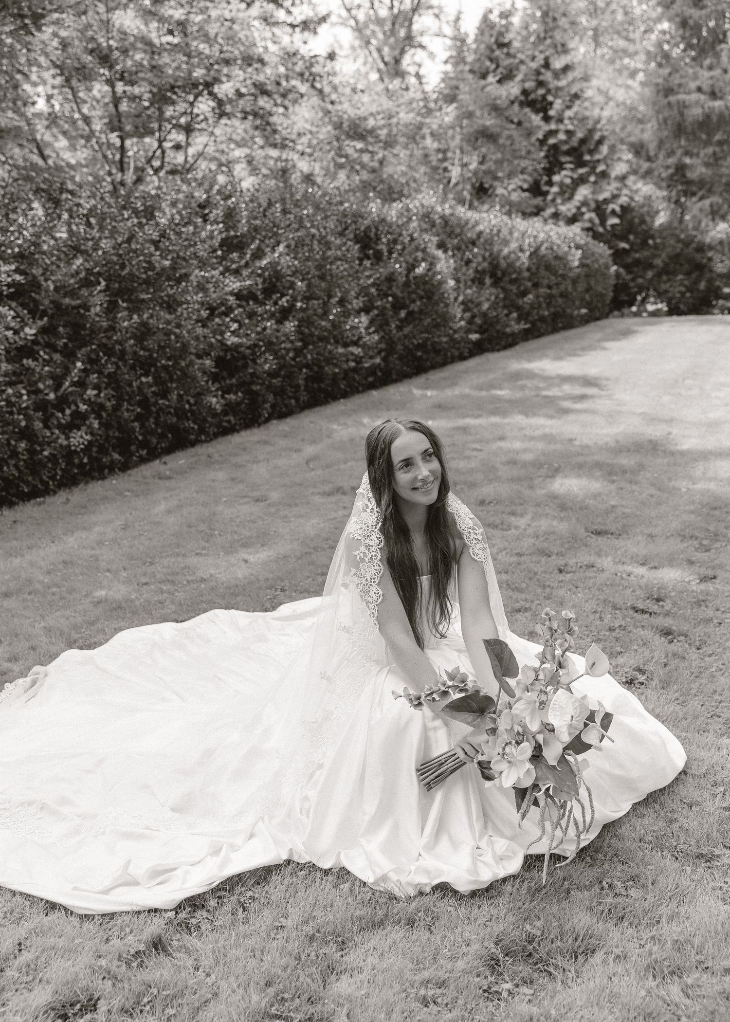 Black and white photo of a bride in a flowing gown and veil sitting on a grass lawn holding a bouquet.