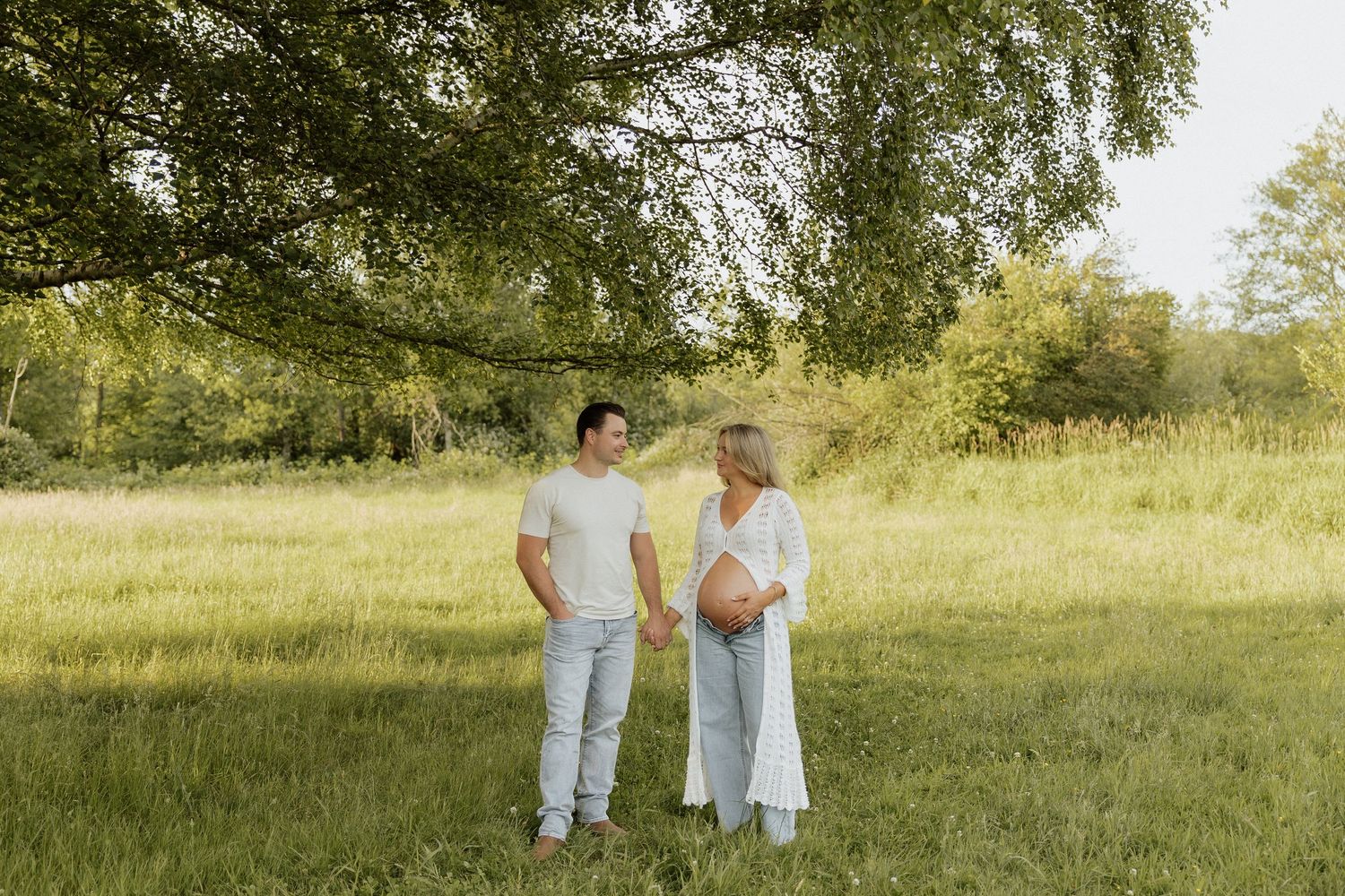Pregnant couple holding hands in a sunny green meadow under a large tree, wearing white and light blue outfits.