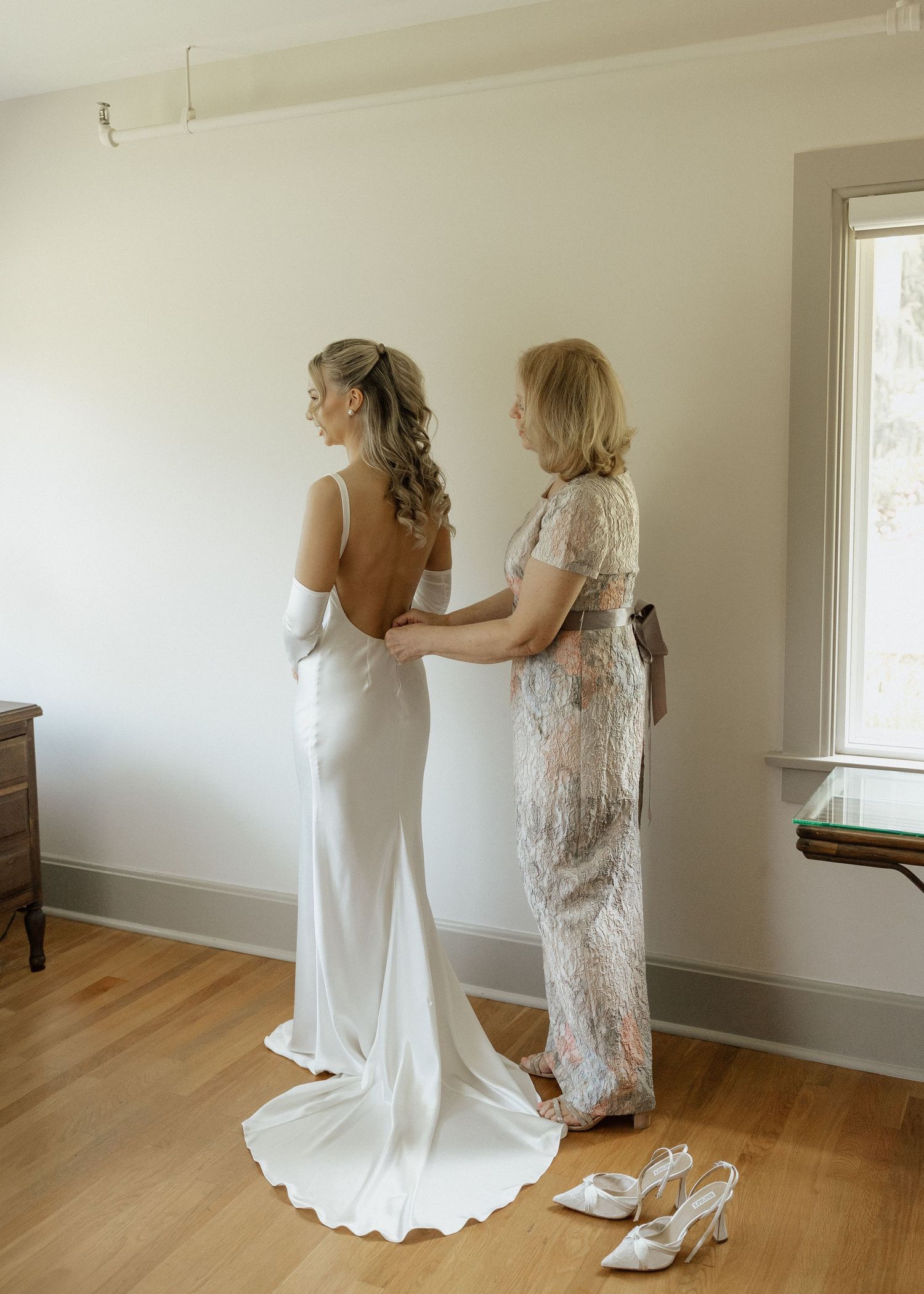 Bride in white gown getting dress fastened by older woman in floral dress, white heels on floor nearby.