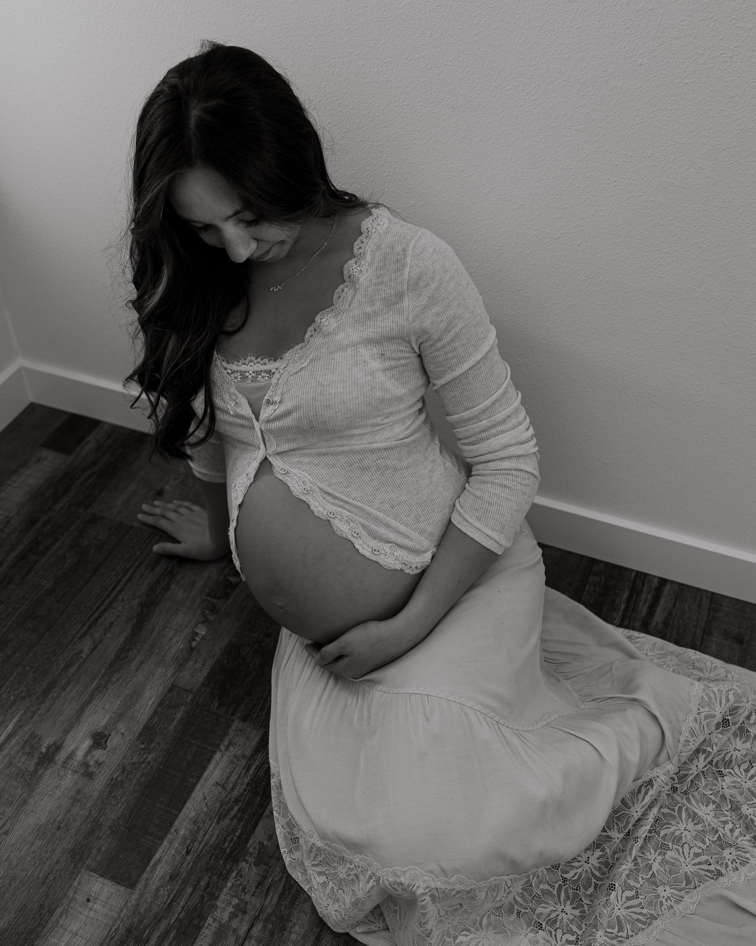 An expectant mother in a white lace dress sits on wooden floor during maternity photoshoot.