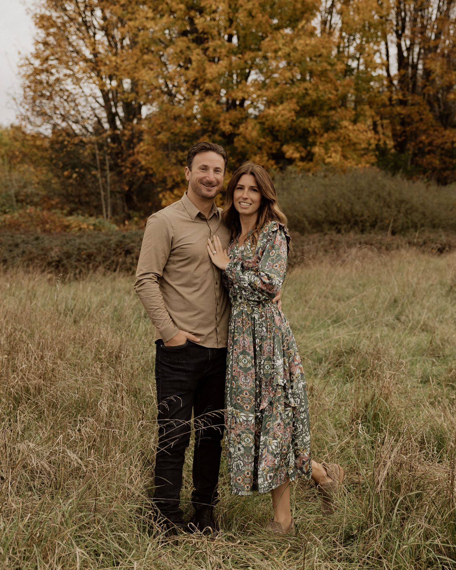Couple posing together in autumn field with golden trees and tall grass.