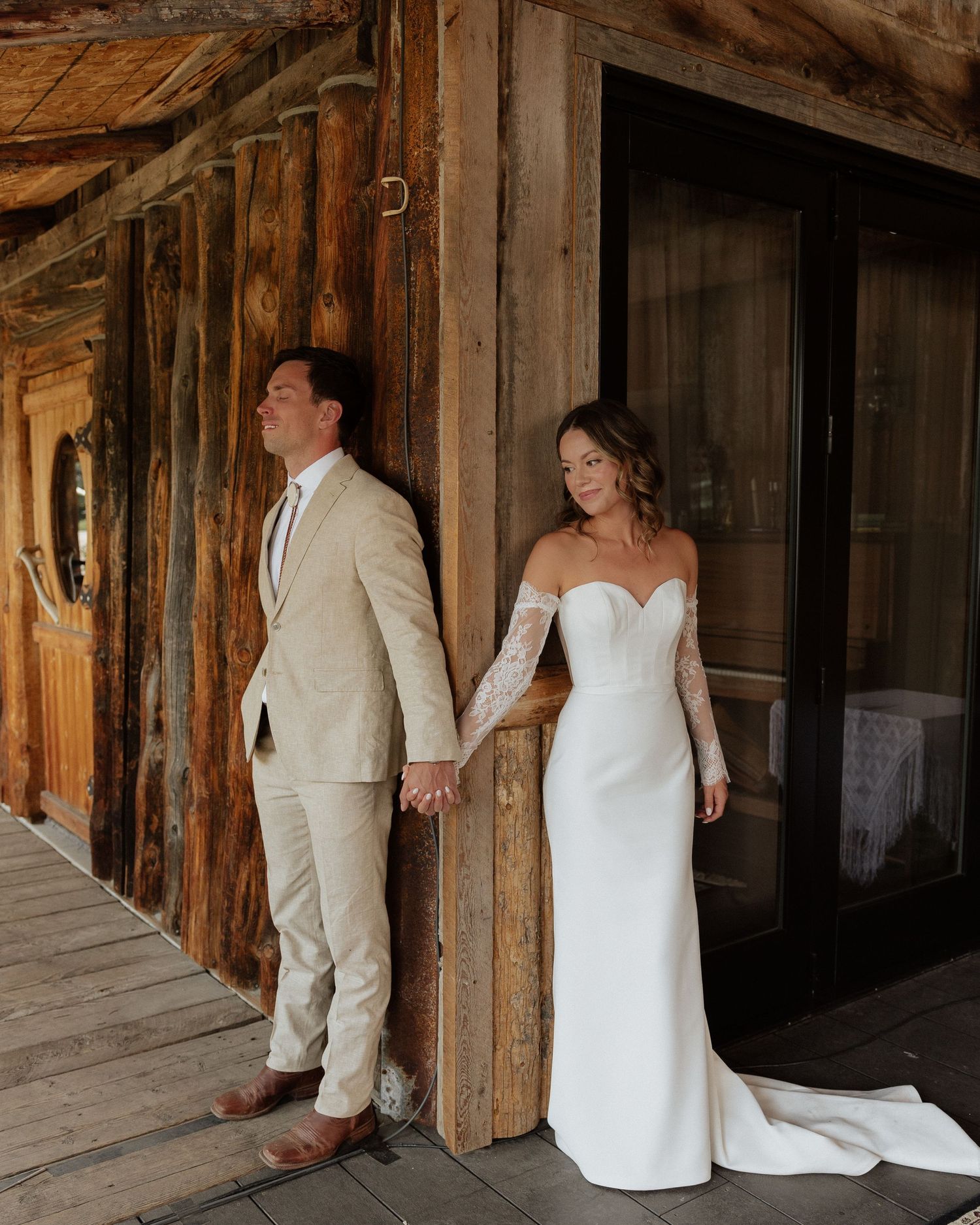 Bride in elegant white gown and groom in tan suit share intimate moment against rustic wooden backdrop.