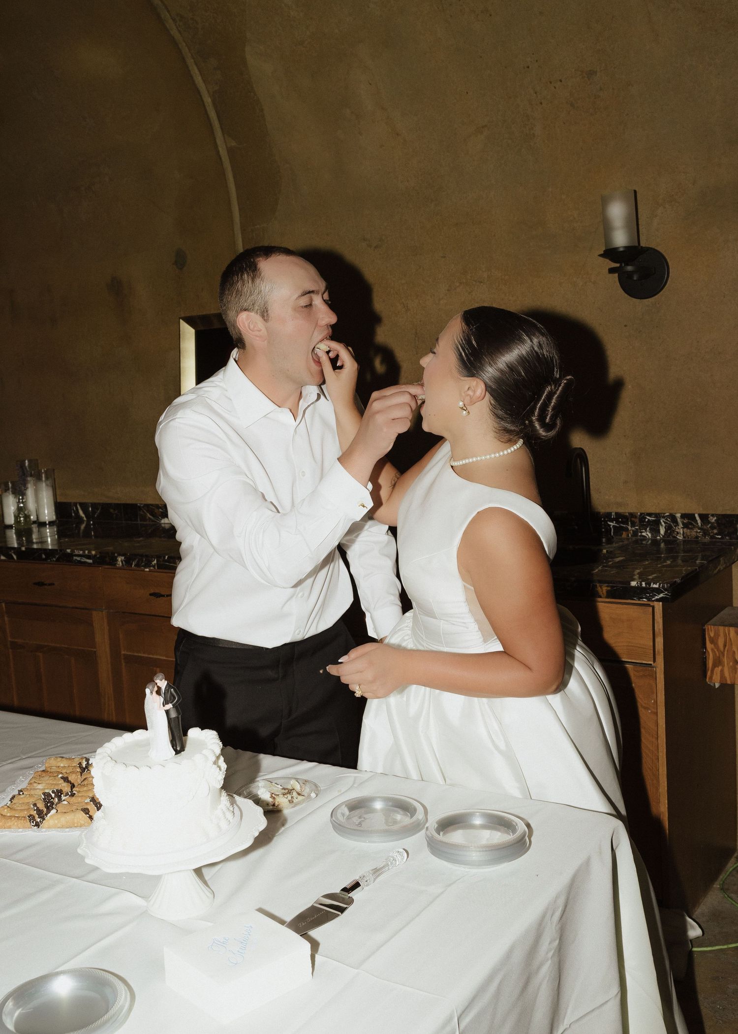Bride and groom feeding each other wedding cake at reception table in rustic venue.