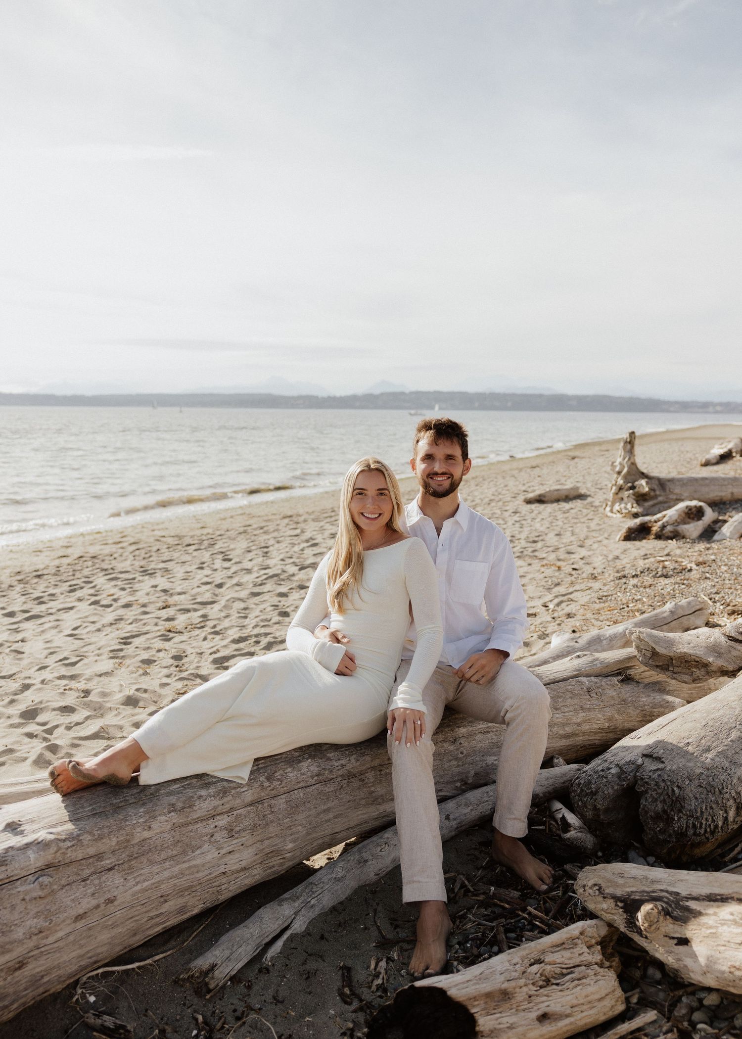A smiling couple in white outfits sits on driftwood on a sandy beach, with calm ocean waves and mountains in the background.