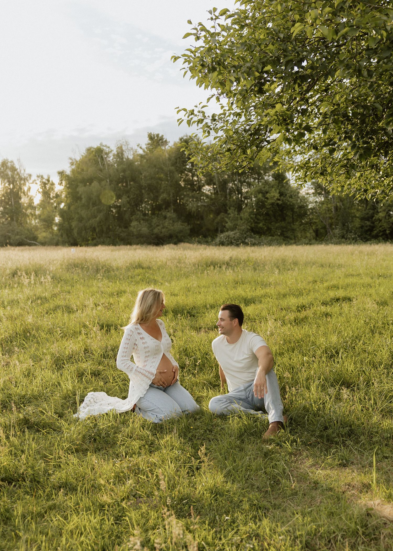 Expecting couple sitting together in a sunlit grassy field, sharing a loving glance at golden hour.