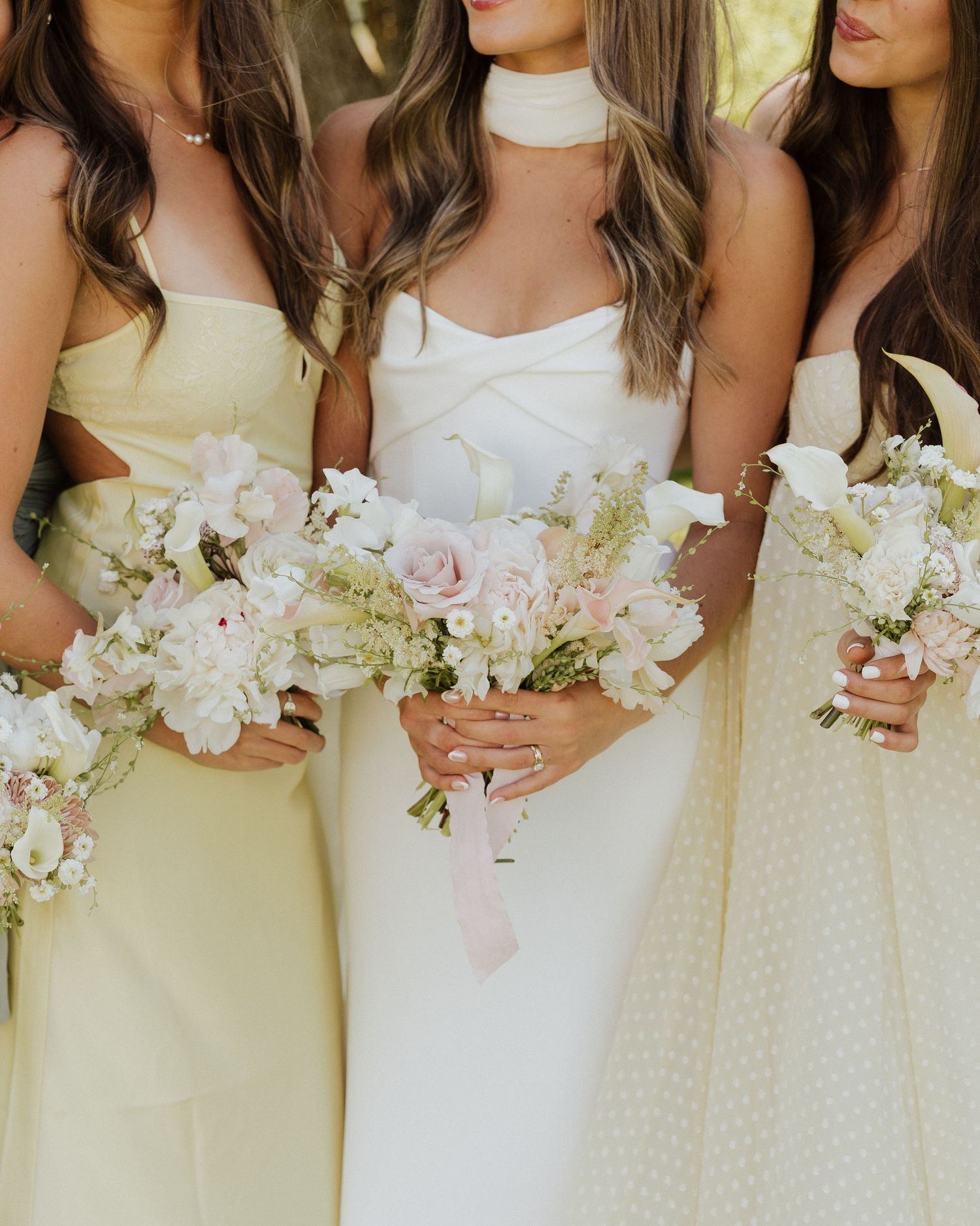Bride in white gown with choker neckline flanked by bridesmaids in pale yellow dresses holding blush and white floral bouquets.