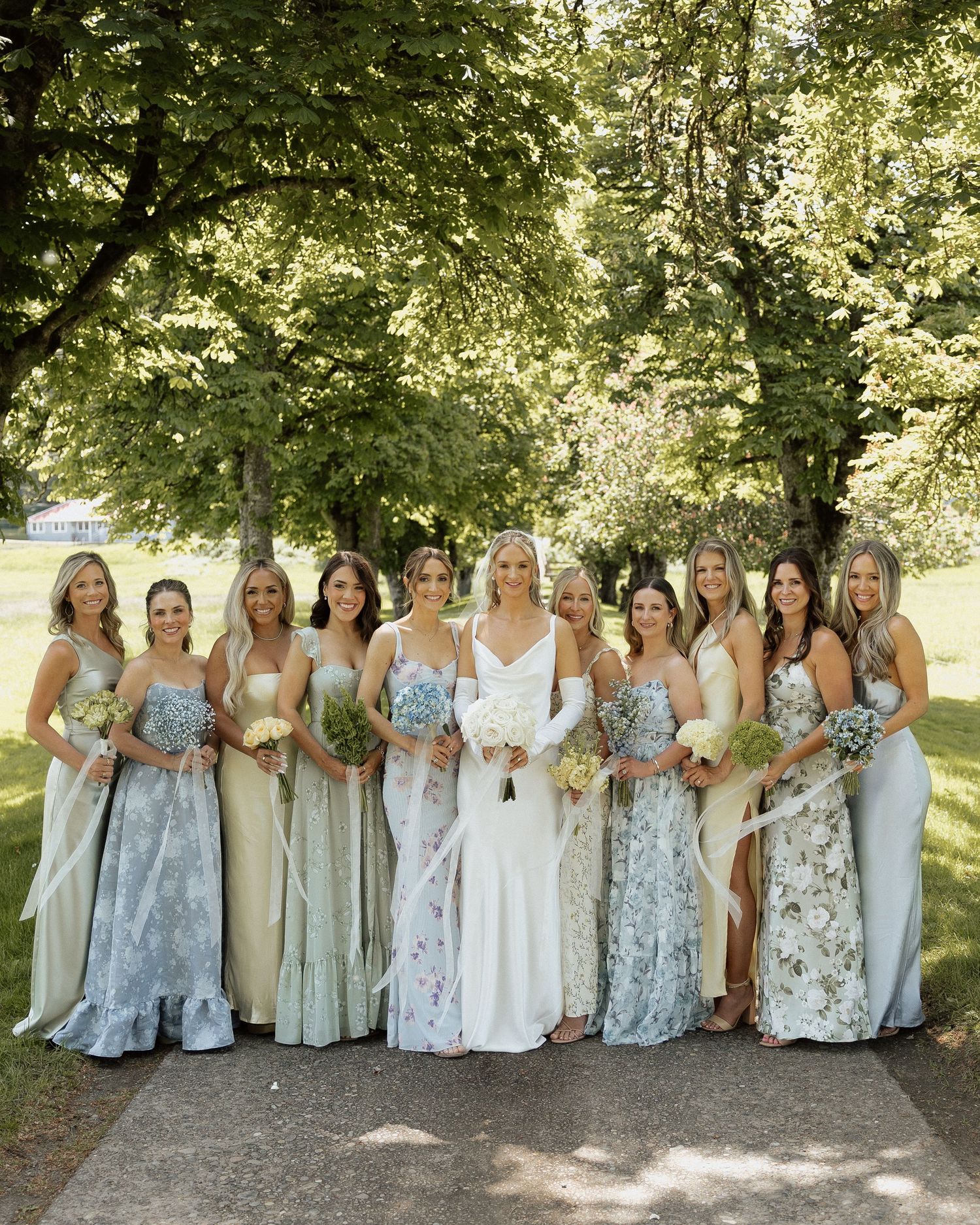 Bride in white gown poses with bridesmaids in mismatched floral and sage dresses outdoors at a garden wedding.