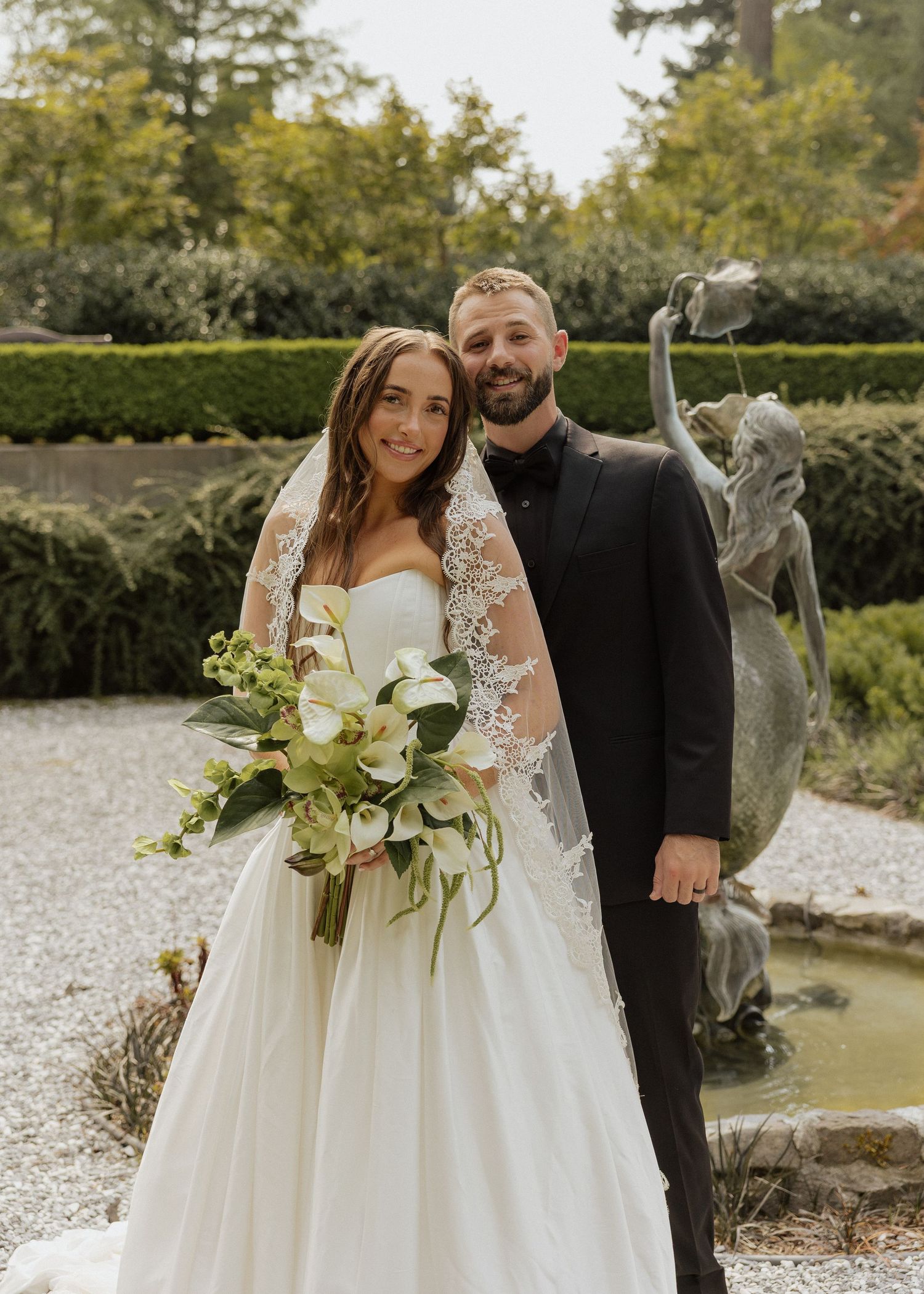 Bride and groom posing together in garden with stone fountain, bride holding white floral bouquet with lace veil.