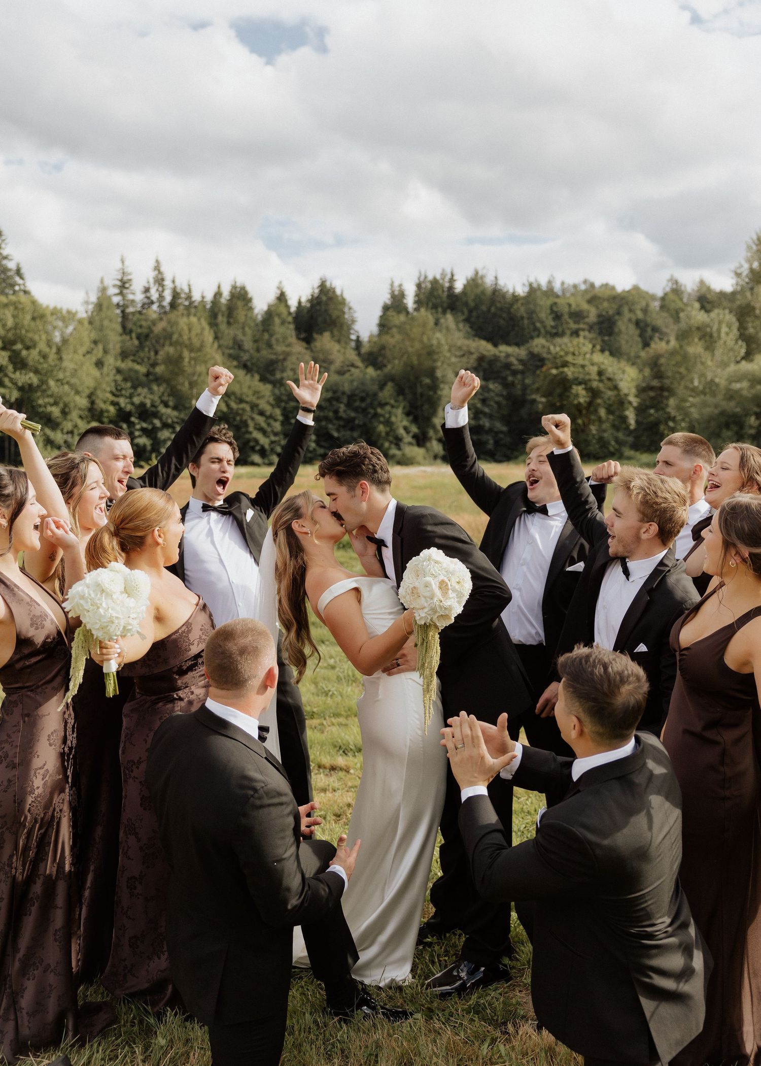 Wedding party cheers as bride and groom share a kiss outdoors, surrounded by lush green trees and overcast sky.