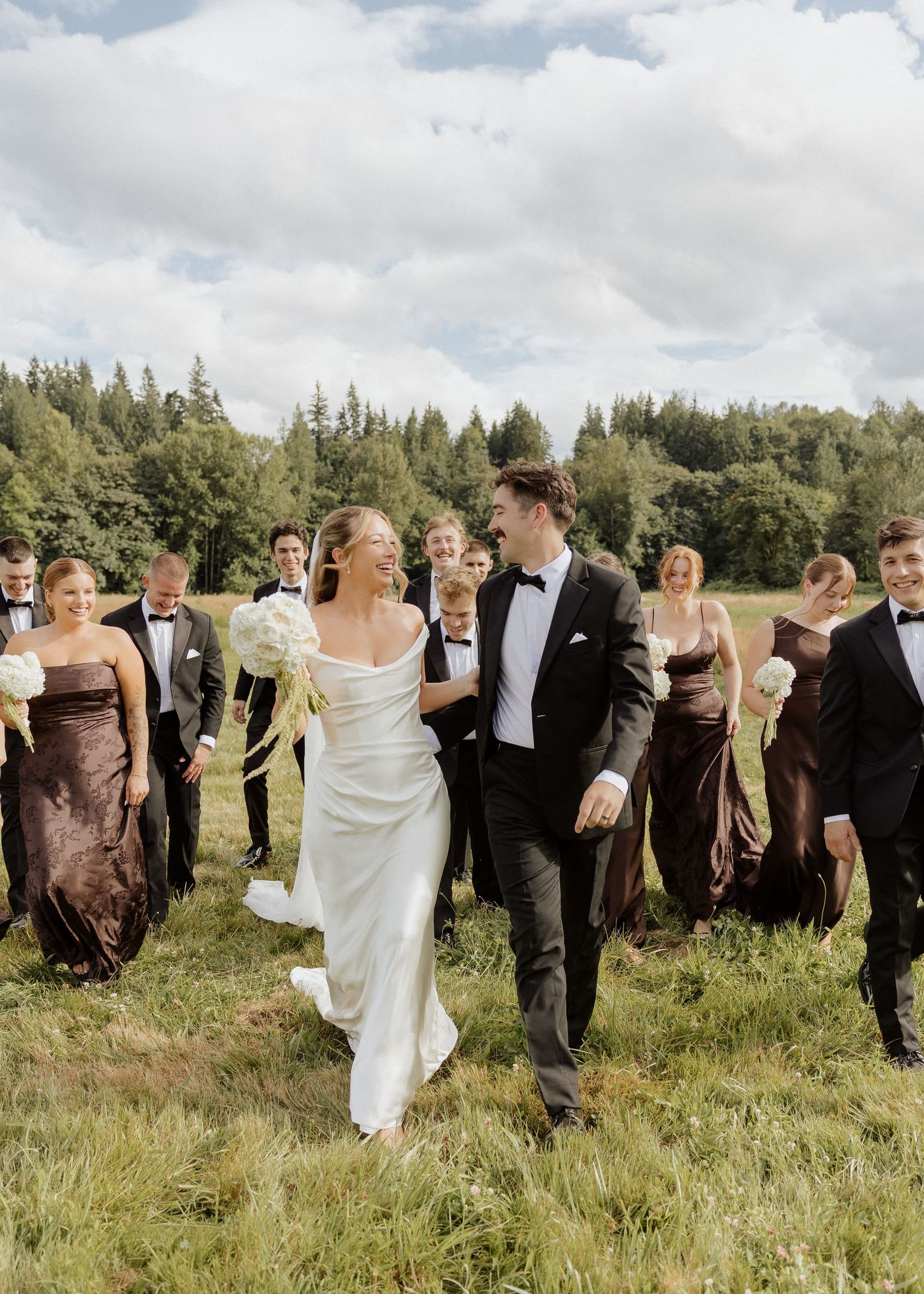 Wedding party walks joyfully through green field, bride in white gown with bridesmaids in brown dresses.