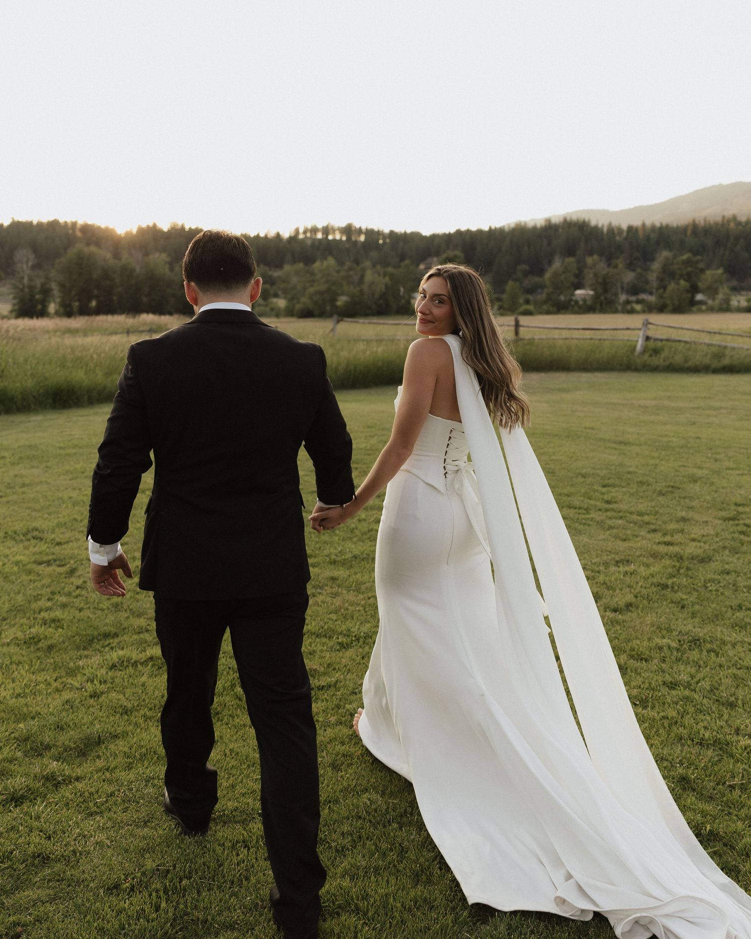 Cropped view of wedding couple walking through a green field with flowing white dress trailing behind.