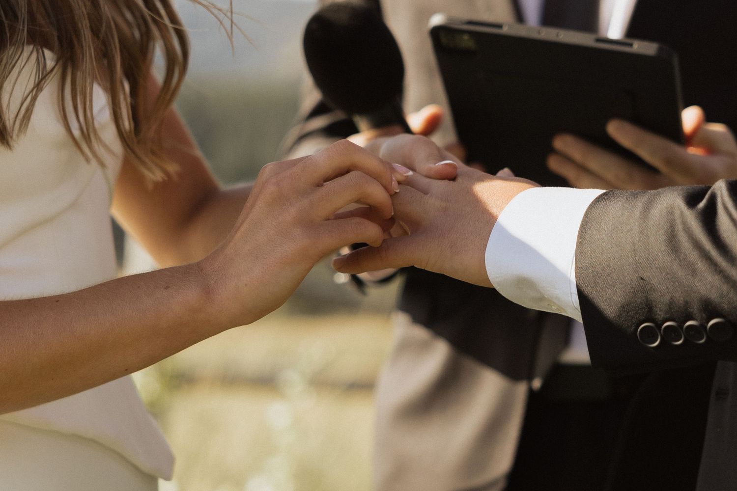Bride placing a wedding ring on groom's finger during outdoor ceremony with officiant holding tablet.