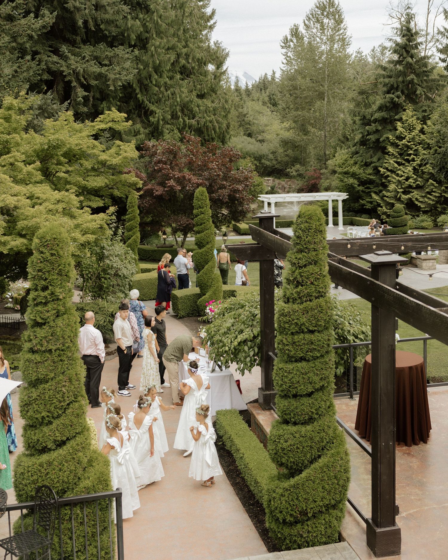 Aerial view of wedding guests and flower girls in white dresses walking through manicured topiary garden at elegant outdoor venue.