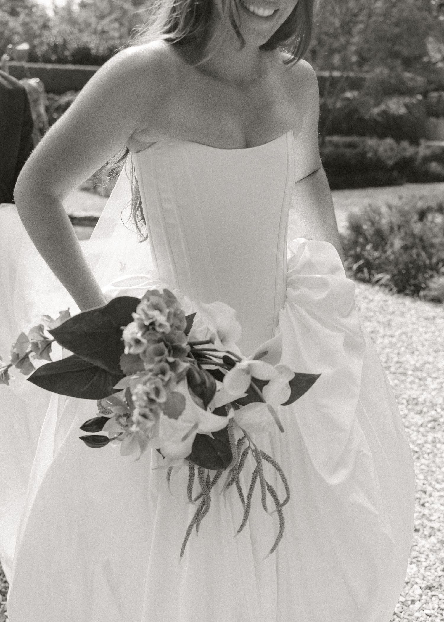 Smiling bride in strapless white wedding gown holding a bouquet of flowers in a black and white outdoor photo.
