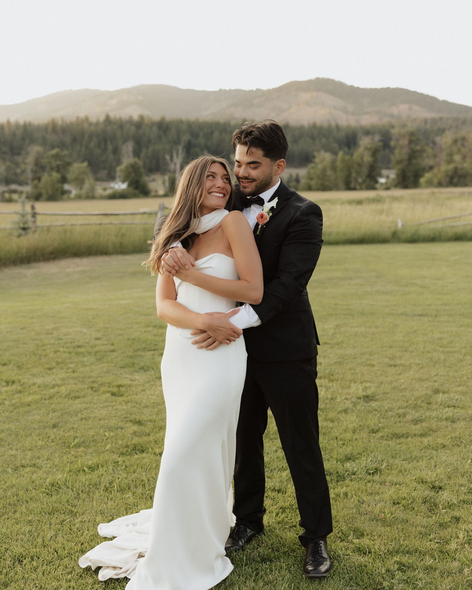 A bride in a white gown and groom in a black tuxedo embrace on a green lawn with mountains in the background.
