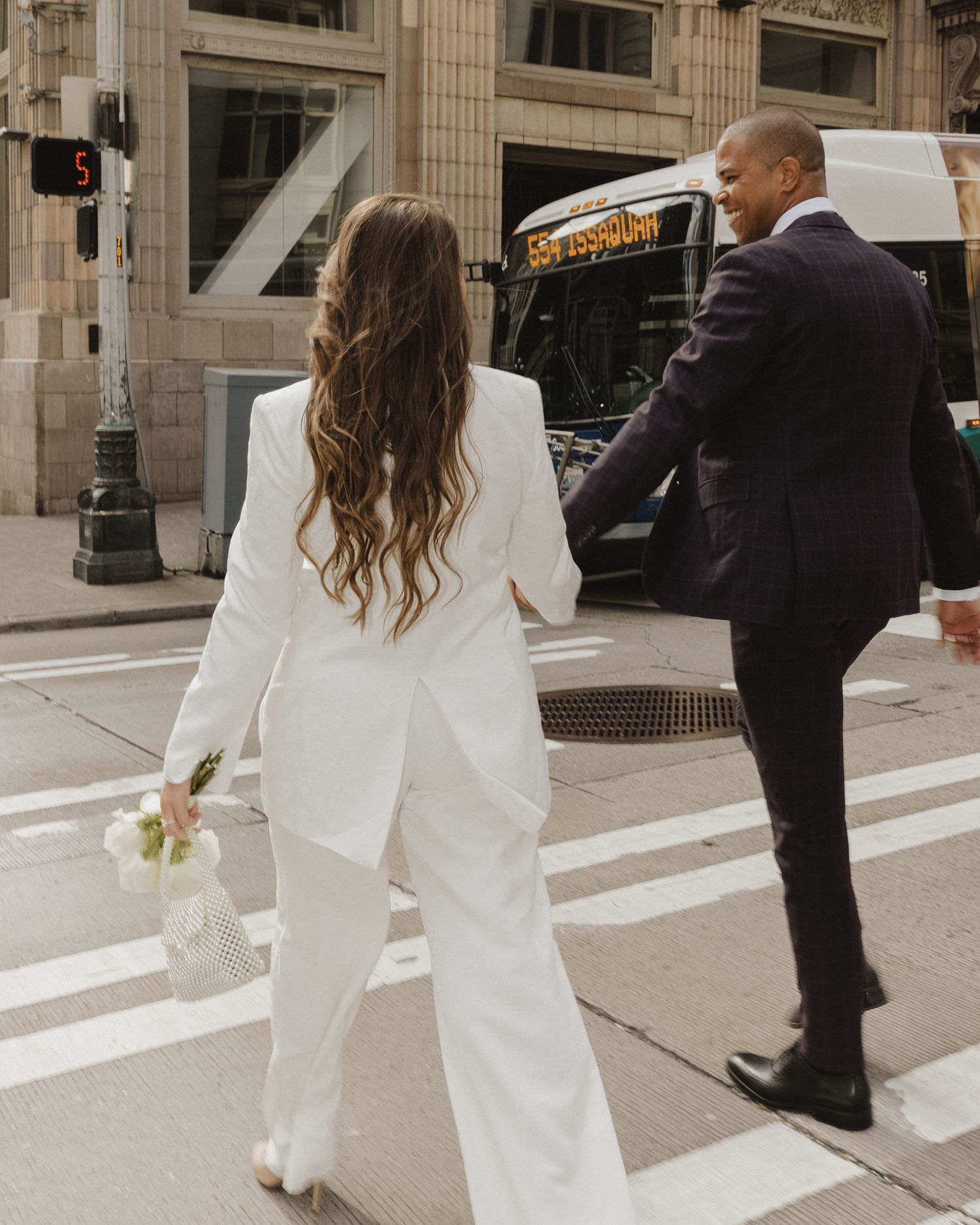 A bride in a white pantsuit holding a bouquet crosses a city street with a groom in a dark suit.