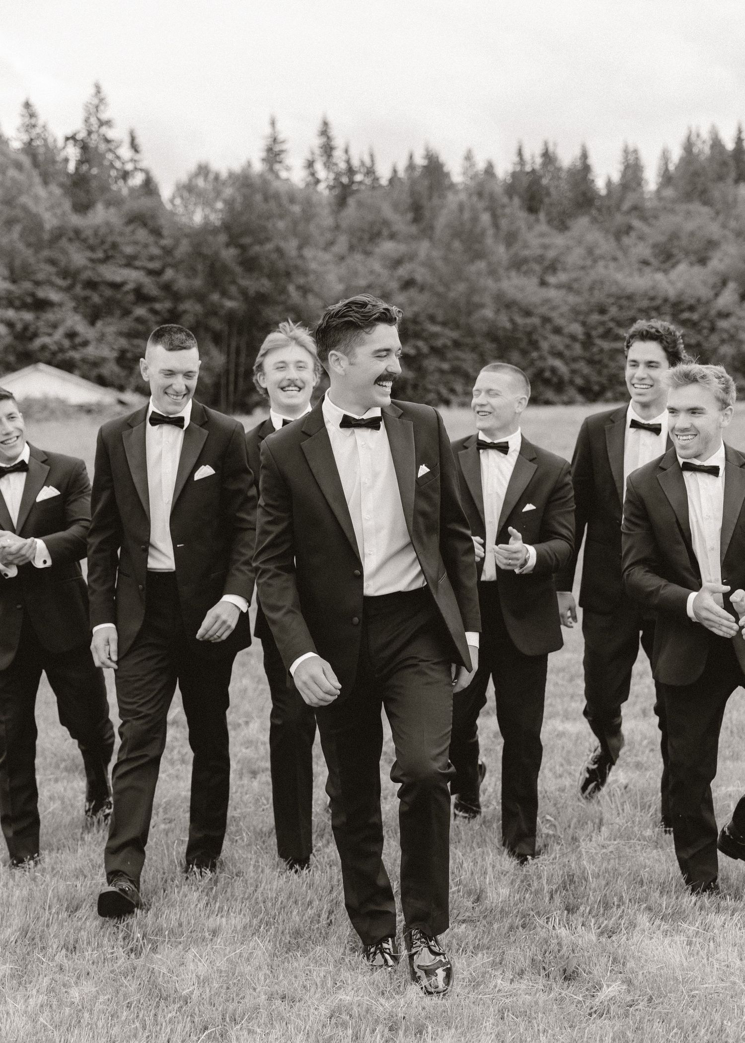 Black and white photo of groom and groomsmen in tuxedos walking outdoors with forest backdrop.