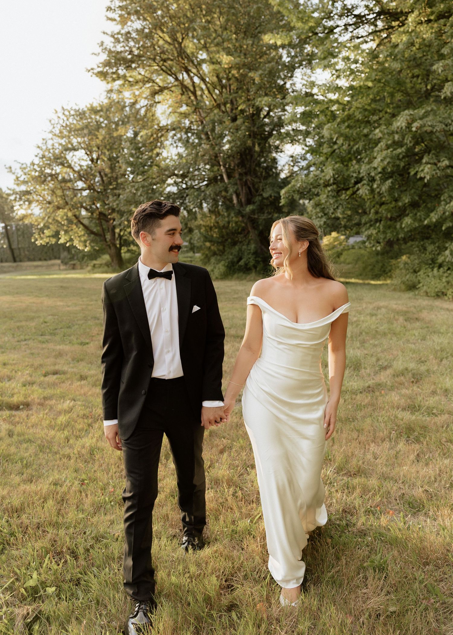 Bride in off-shoulder white gown and groom in black tuxedo walking hand in hand in field.