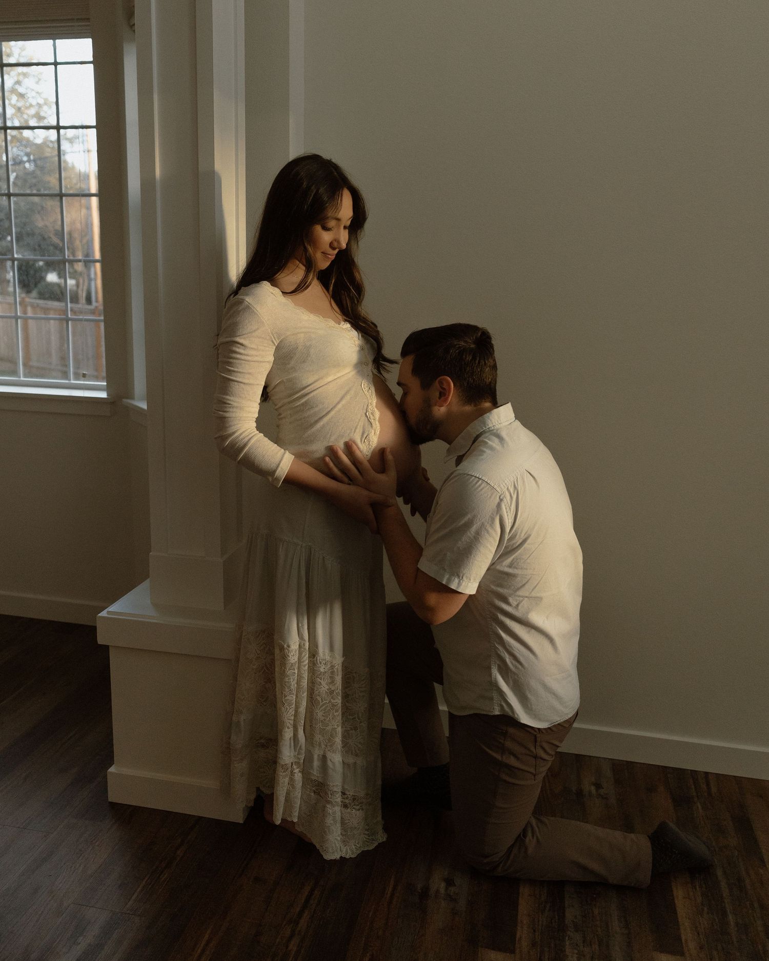 Expecting parents sharing tender moment by window during indoor maternity session.