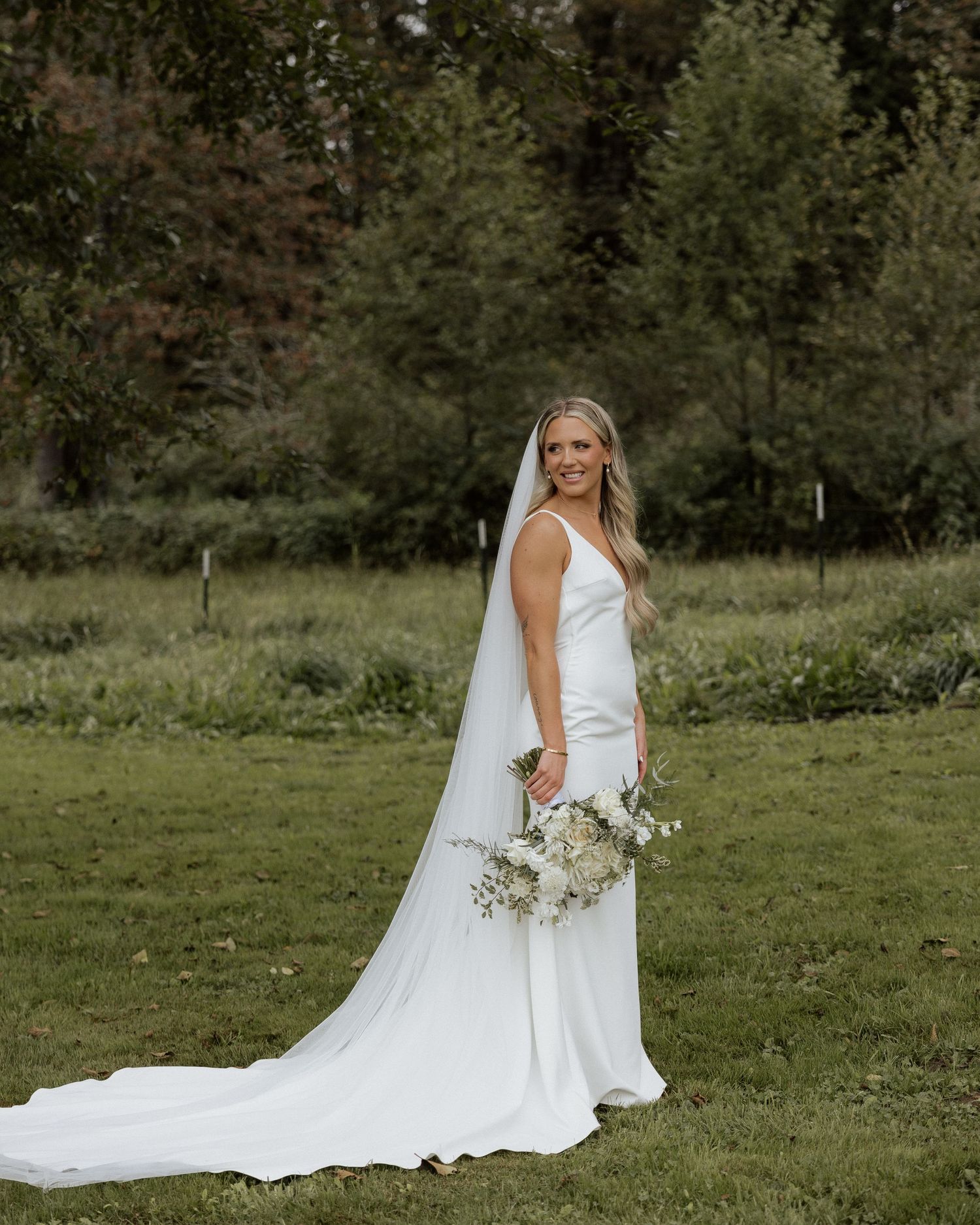 Bride in minimalist white dress poses with bouquet against forest backdrop.