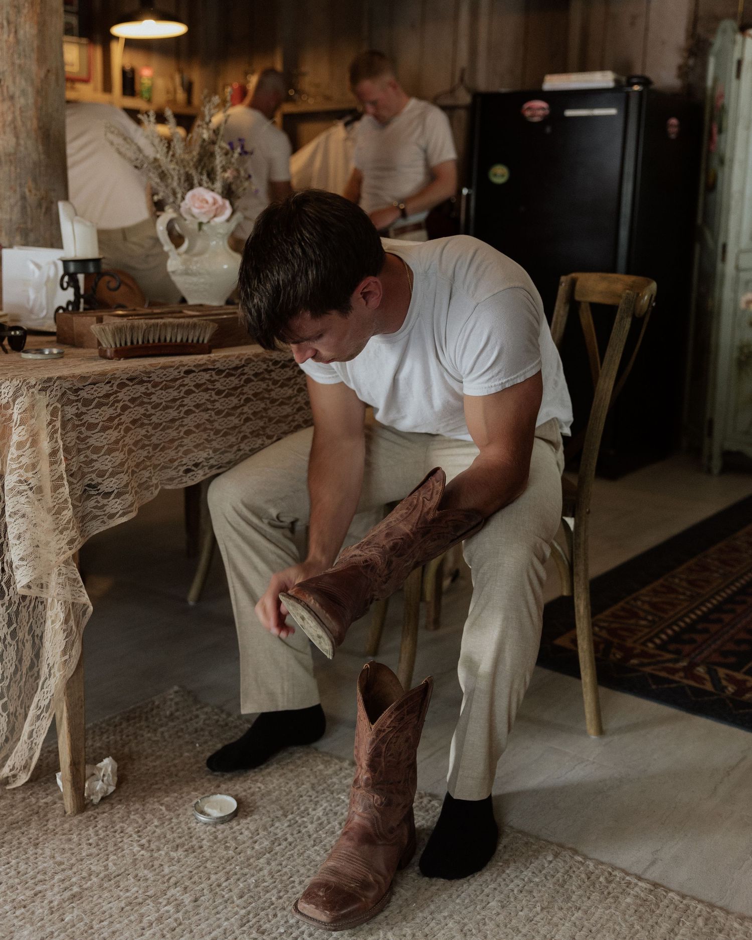 Person in white clothing cleaning brown leather boots while seated on a chair.