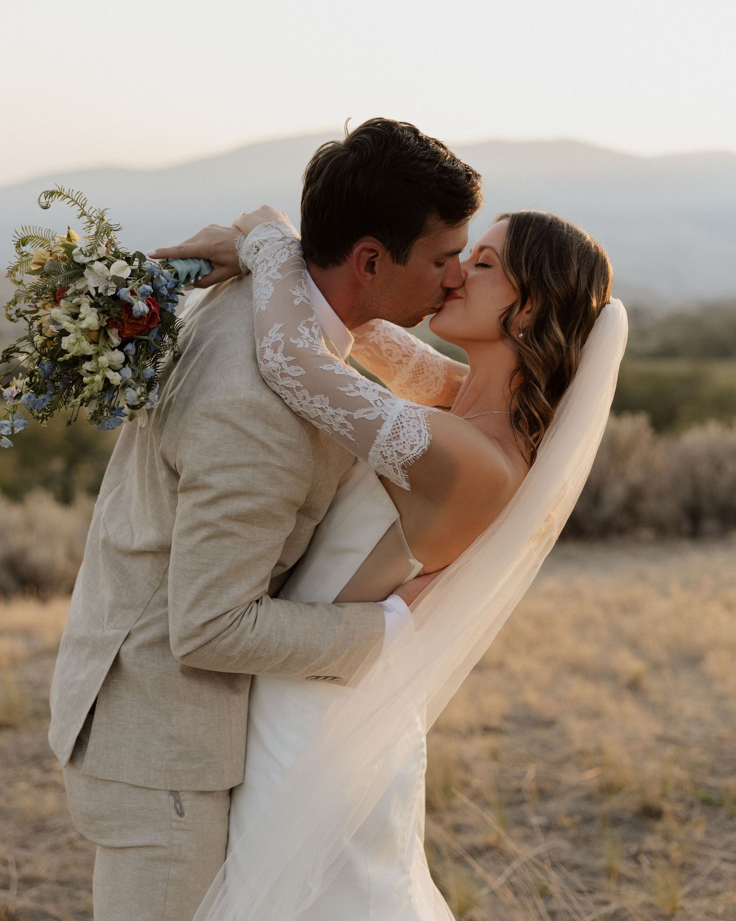 Passionate wedding kiss captured during sunset with mountain landscape and flowing veil.