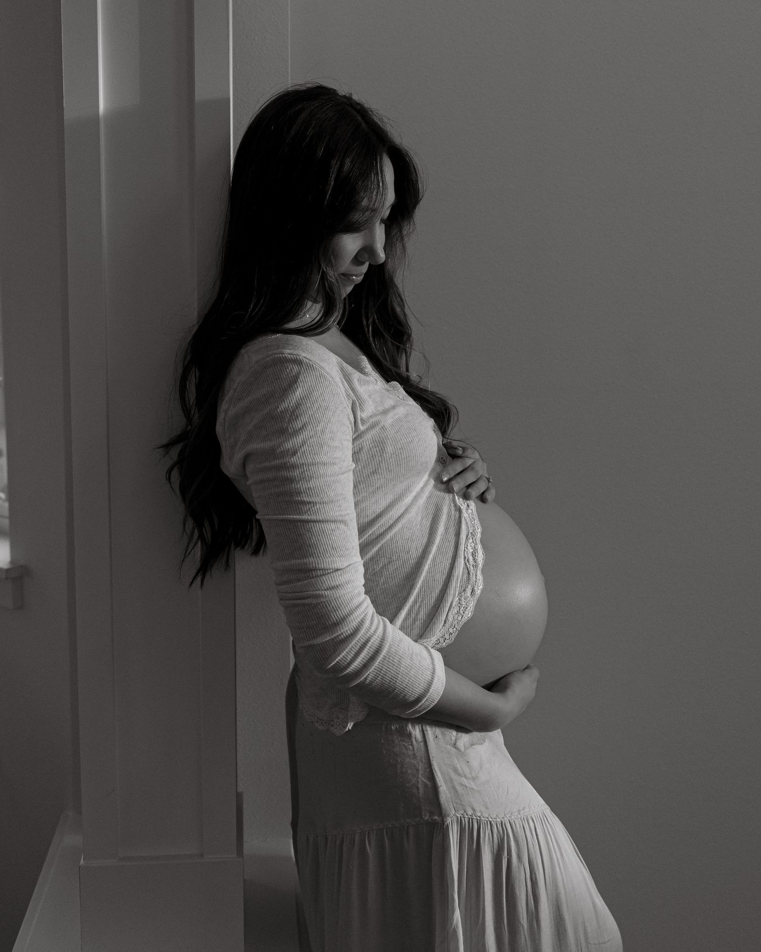 A black and white maternity portrait showing a gentle moment by a window.