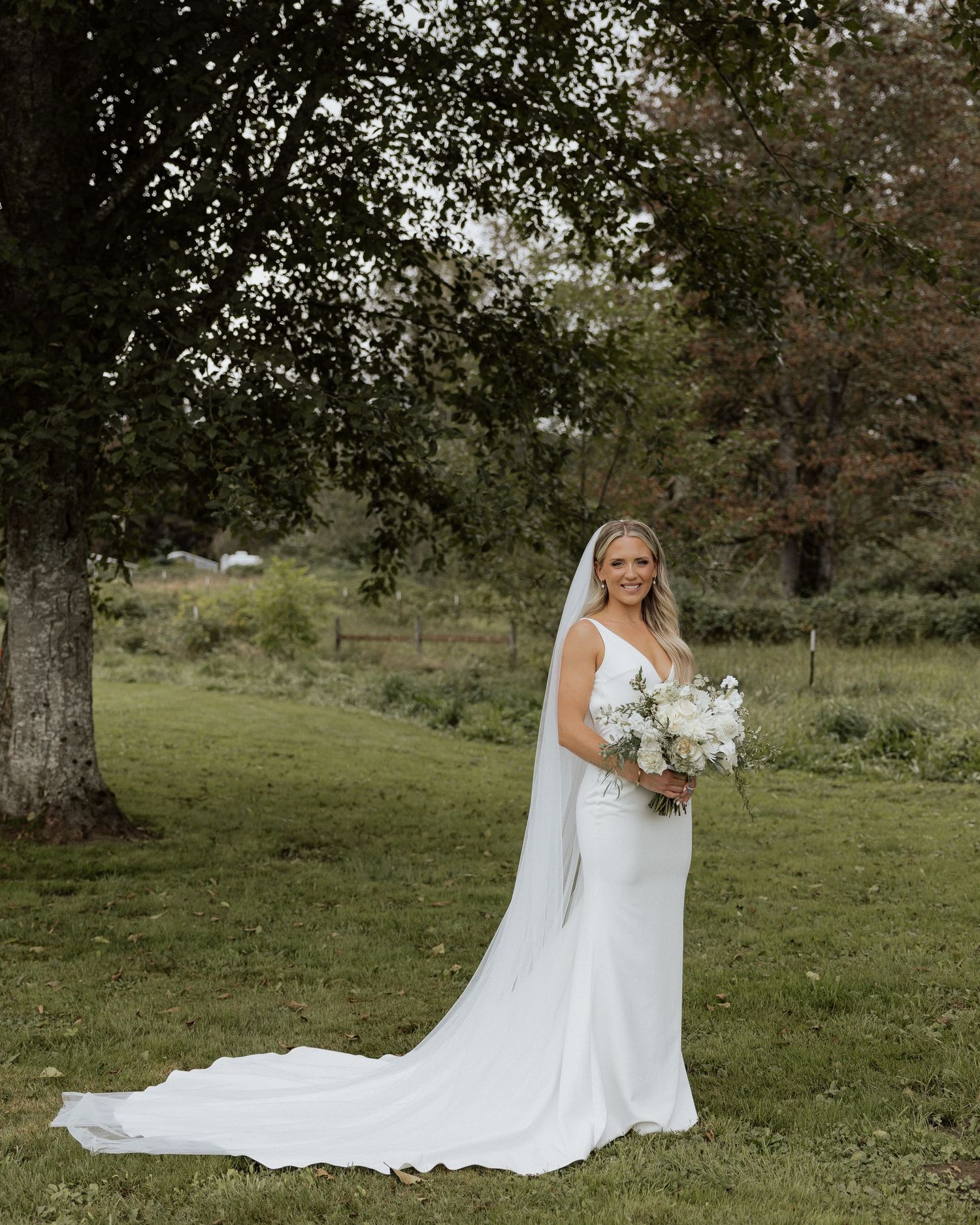 Bride in elegant white gown with train poses with bouquet in pastoral setting.