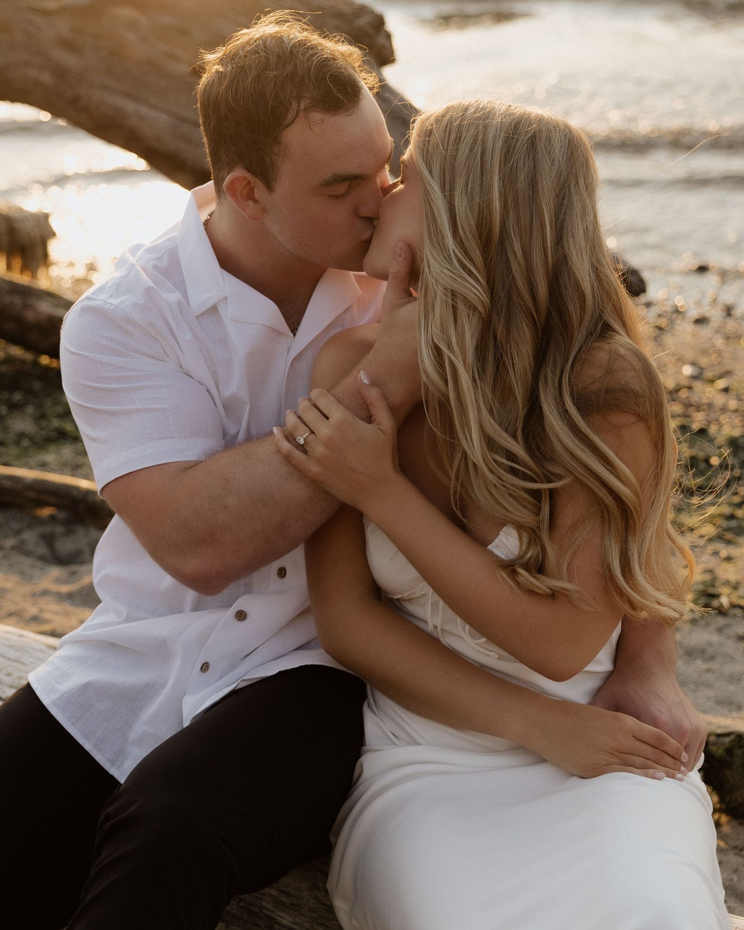 Intimate couple in white clothing sharing a tender kiss on a beach at golden hour.