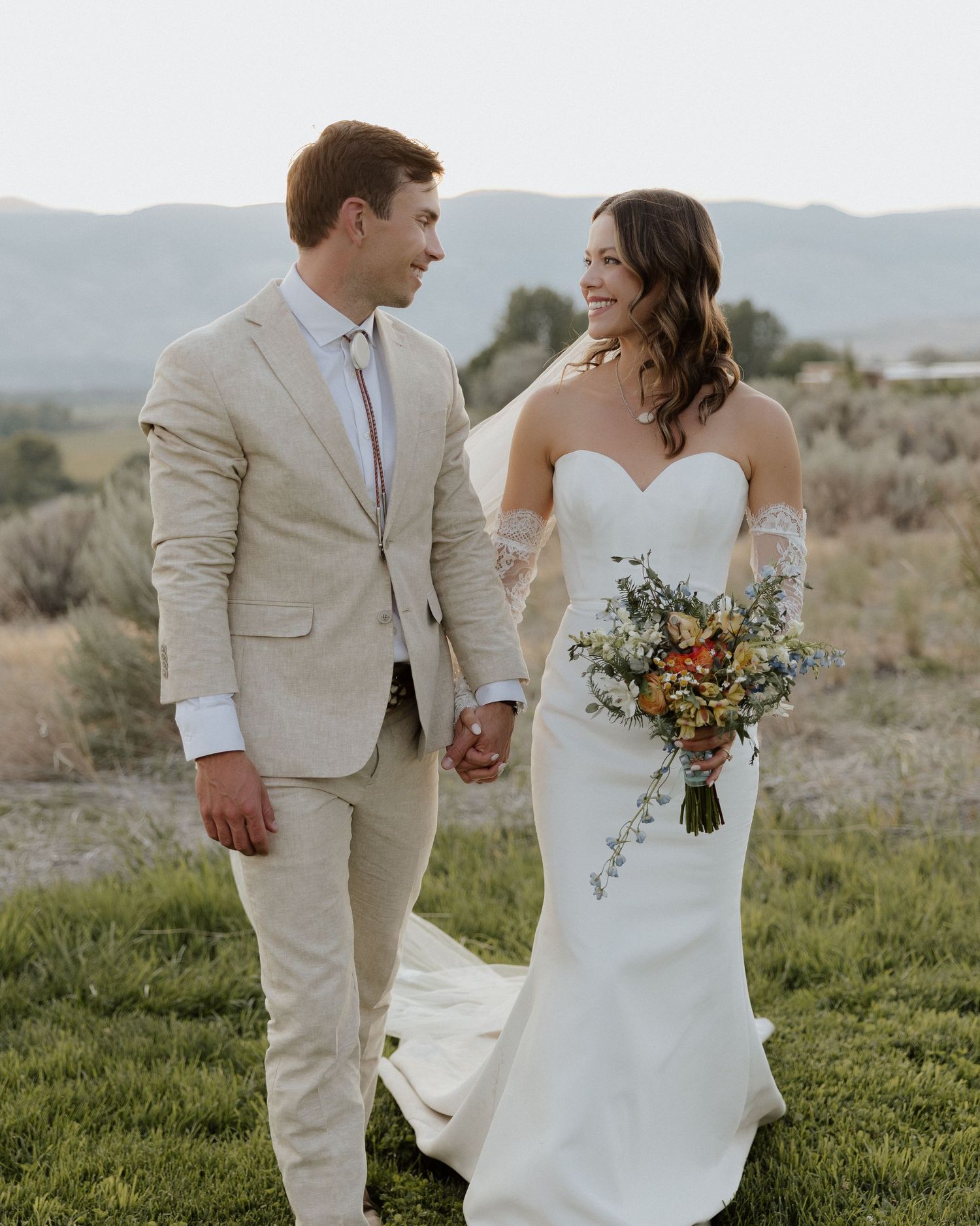 Happy newlyweds walk hand in hand on mountain meadow at sunset.