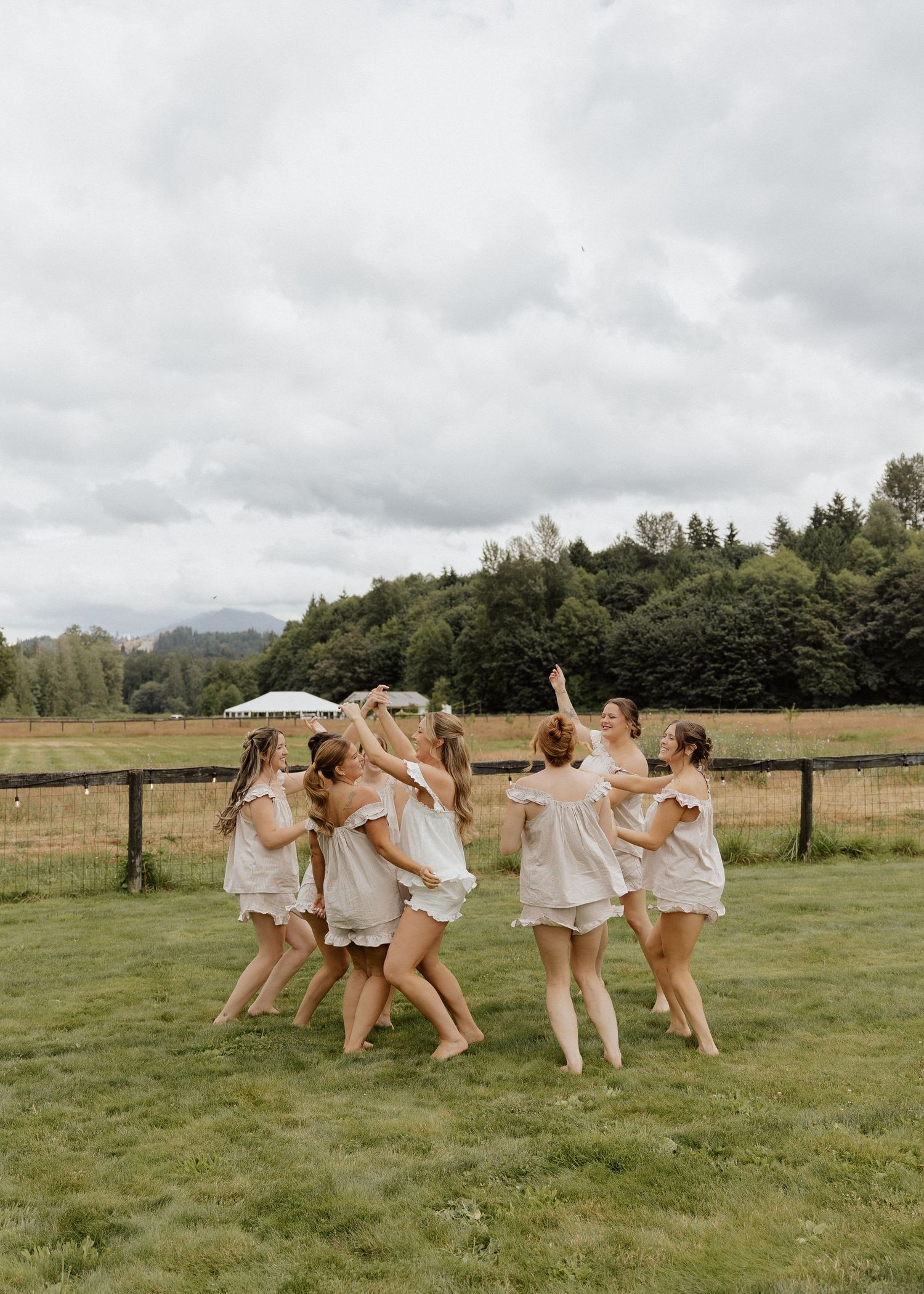 A group of bridesmaids in matching white pajamas dancing and celebrating together on a green farm field.