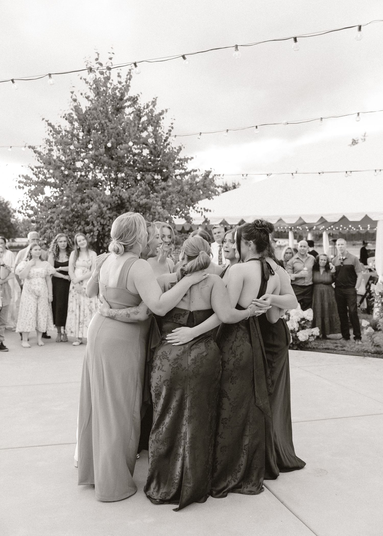 Black and white photo of bridesmaids in long gowns hugging outdoors at wedding reception.