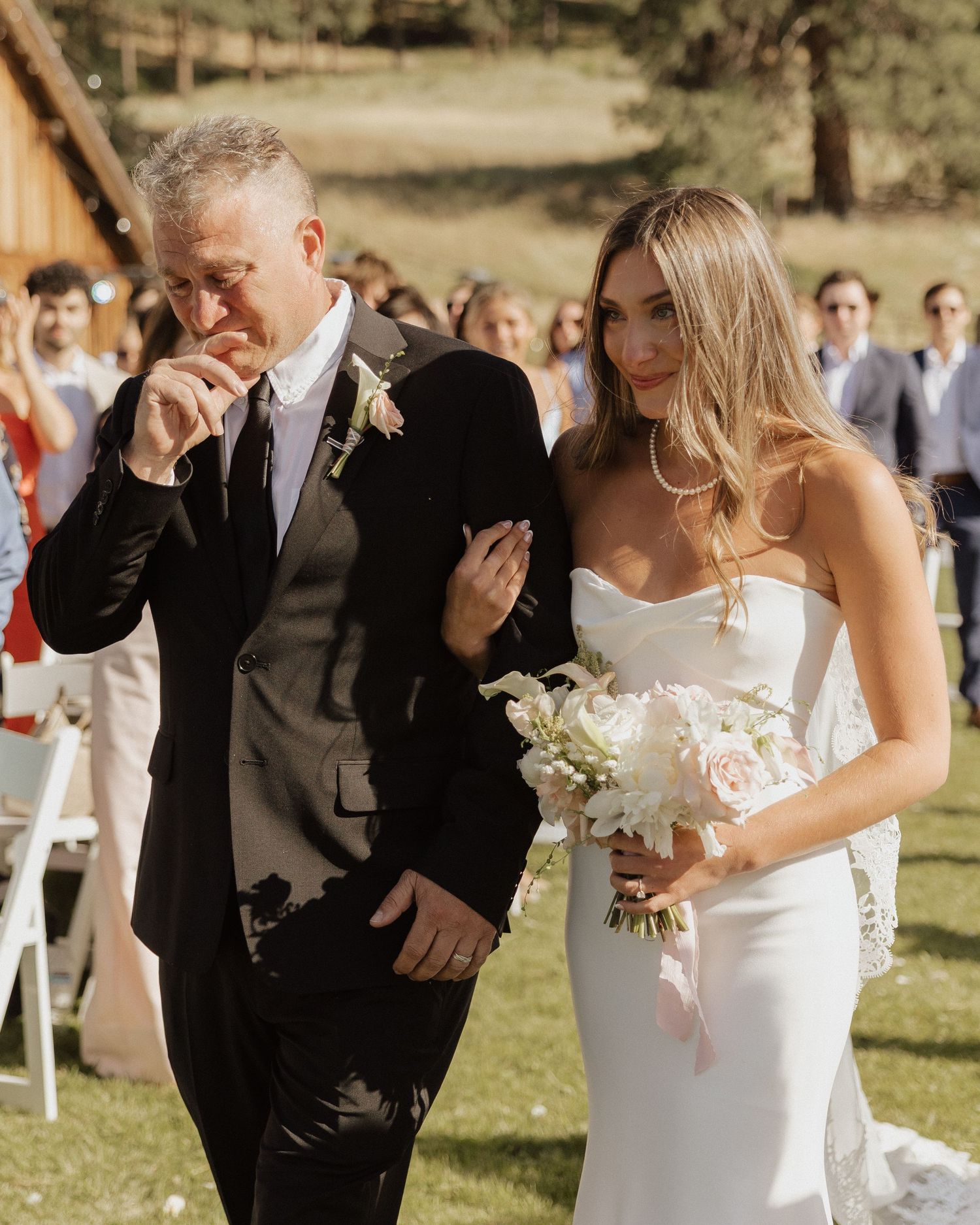 Emotional father walks bride in white gown down outdoor aisle holding bouquet at wedding ceremony.