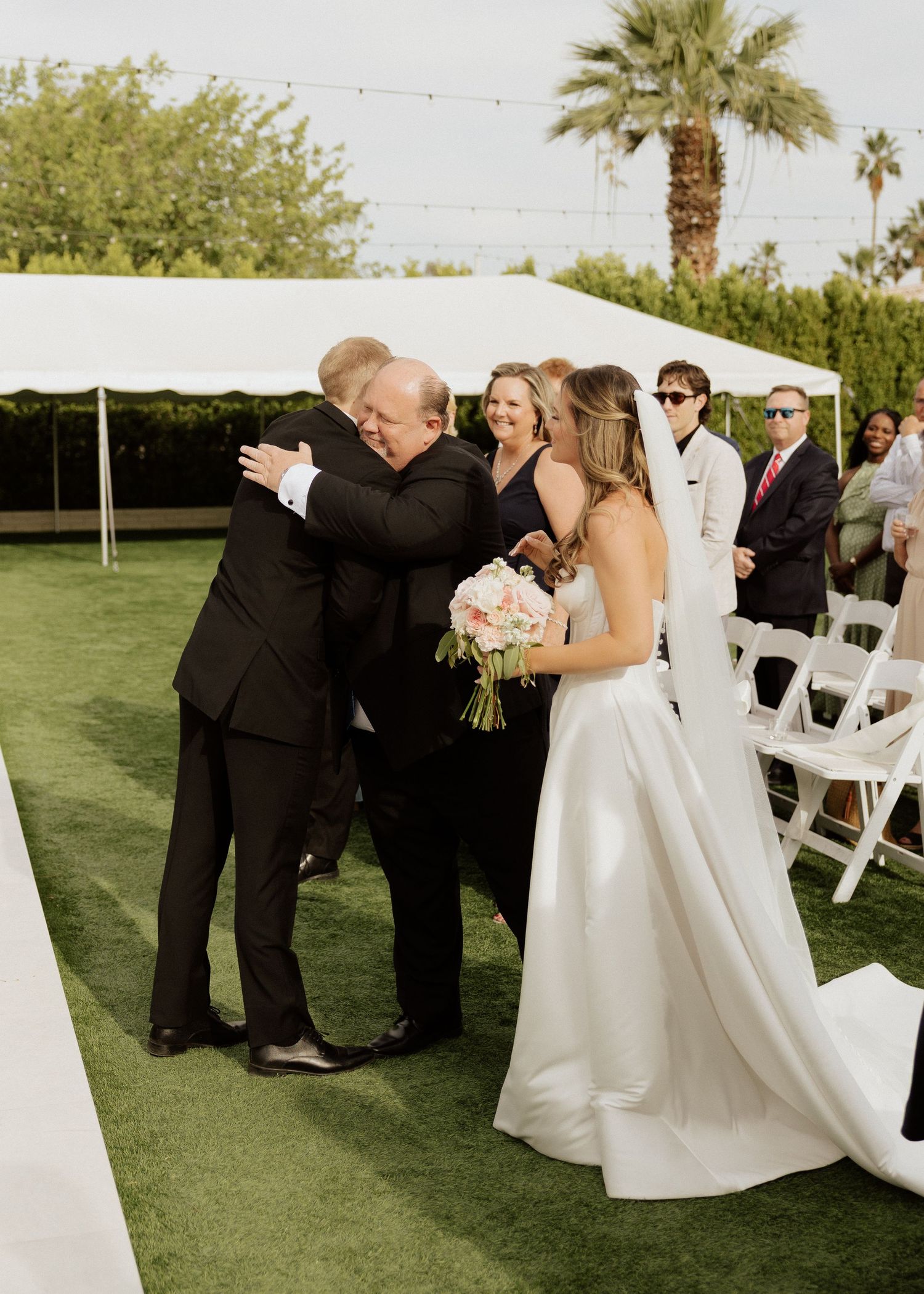Father hugging groom at outdoor wedding ceremony while bride holds pink bouquet under white tent.
