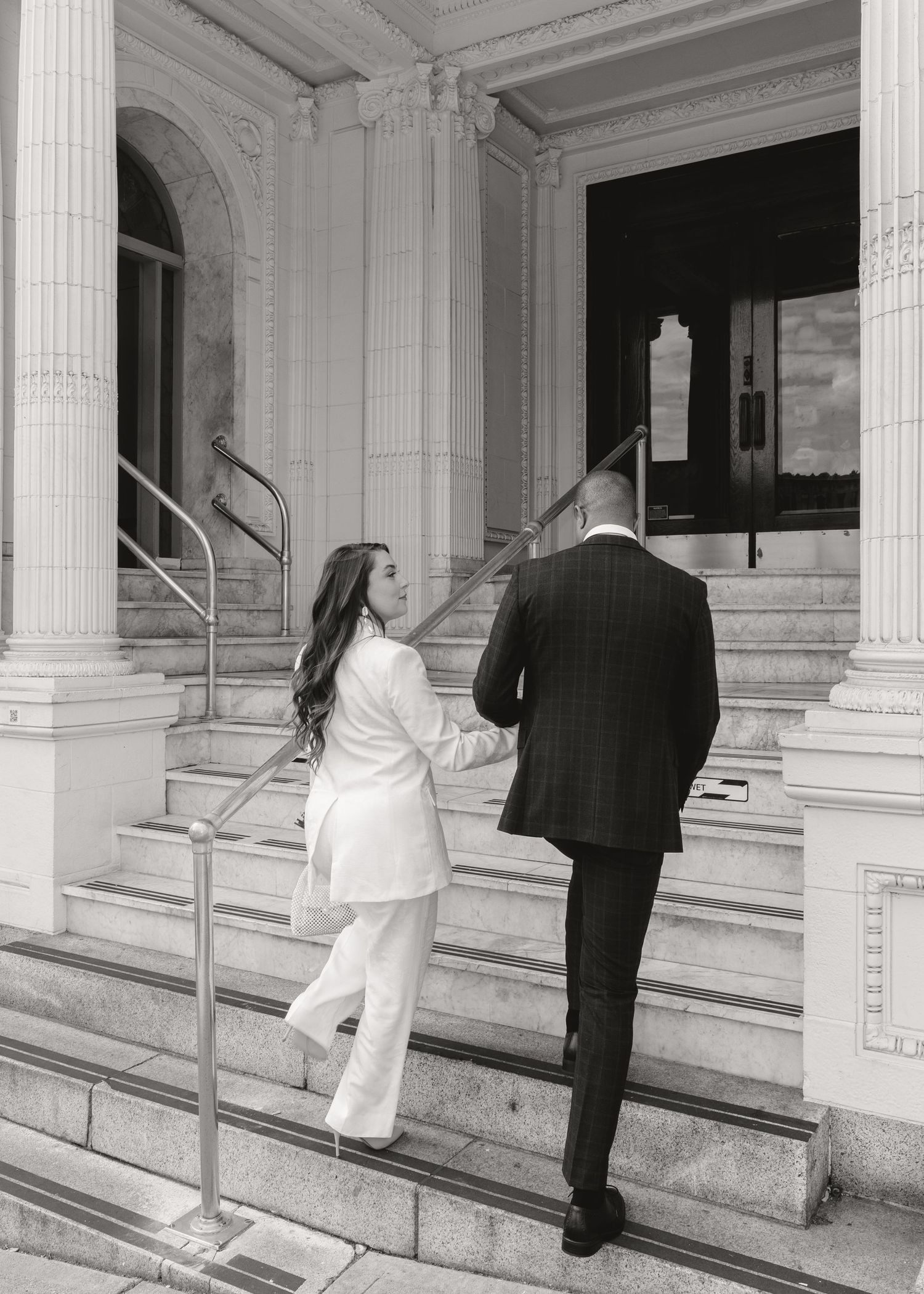 Black and white photo of a couple holding hands walking up grand stone steps of a classical building.