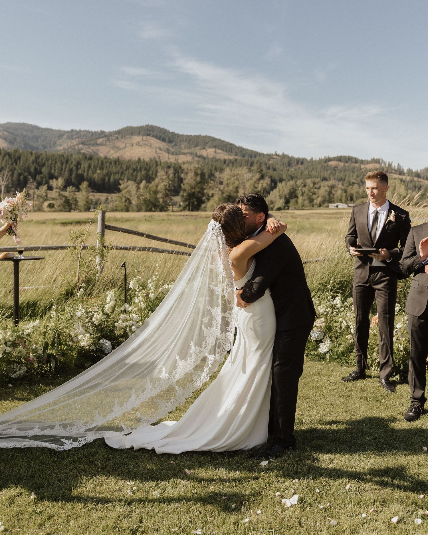 A couple shares their first kiss as newlyweds during an outdoor mountain wedding ceremony with a flowing veil.