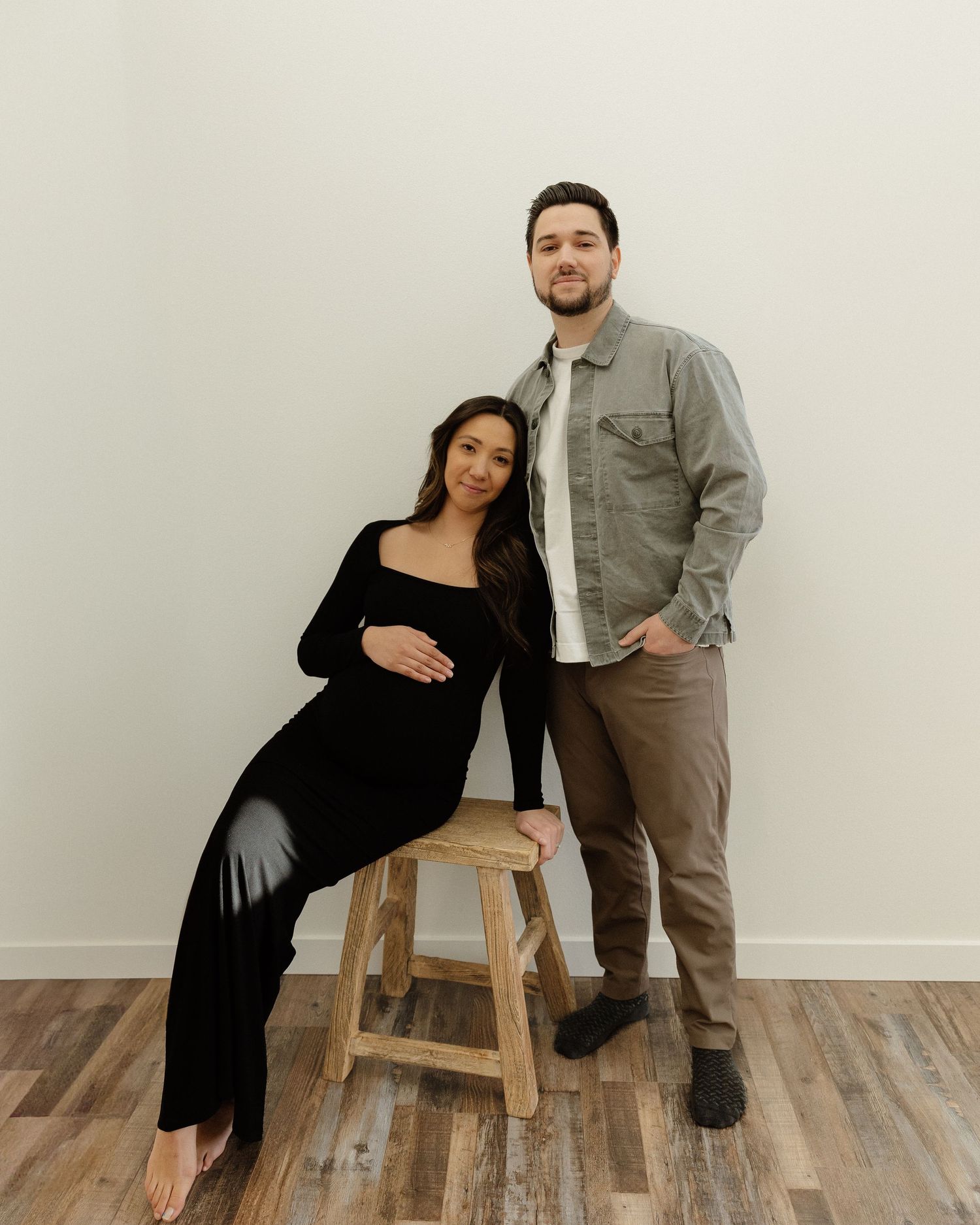 Casual maternity portrait of couple posing with wooden stool against minimalist backdrop.