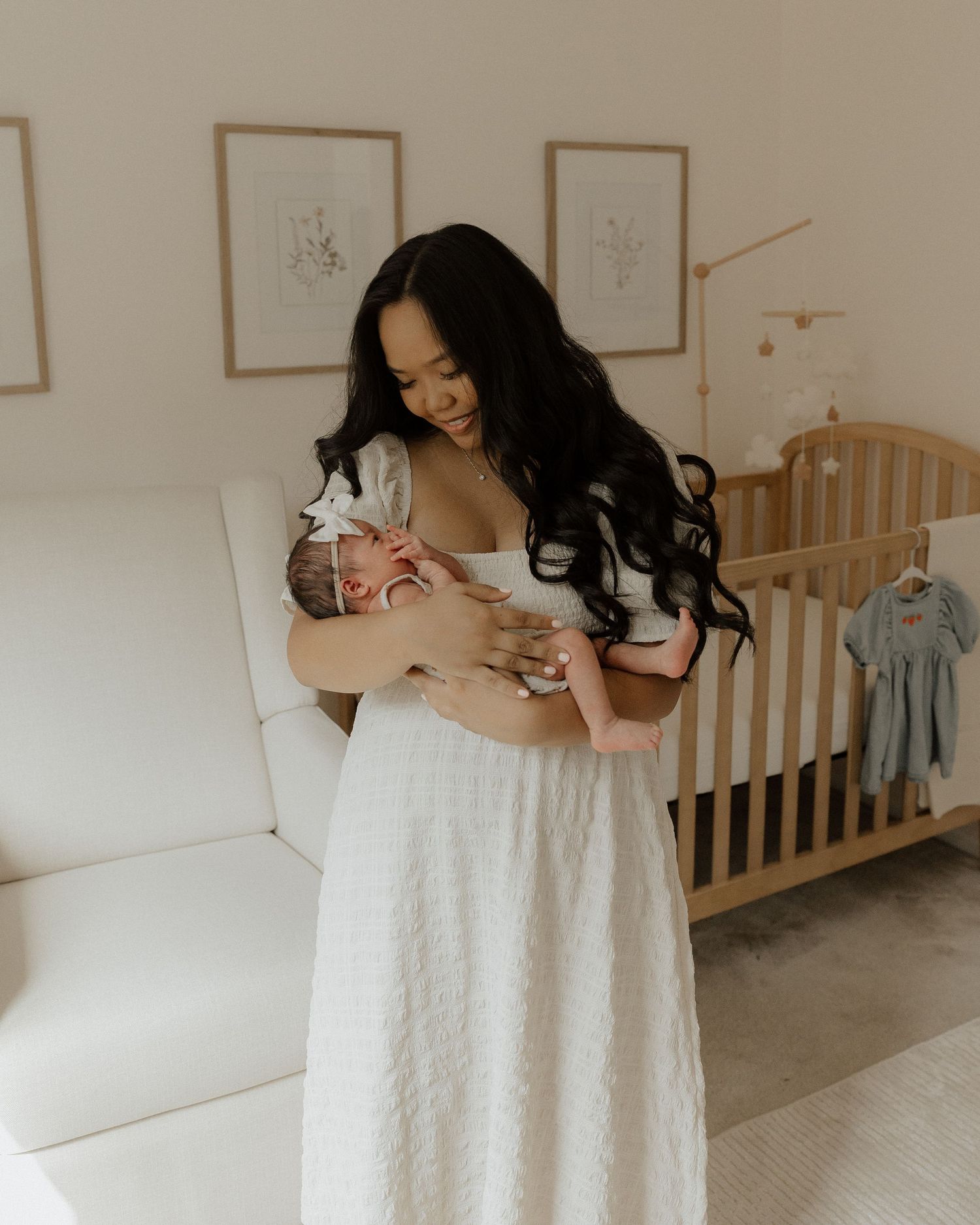 Mother in white dress holding sleeping newborn in bright, modern nursery with wooden crib.