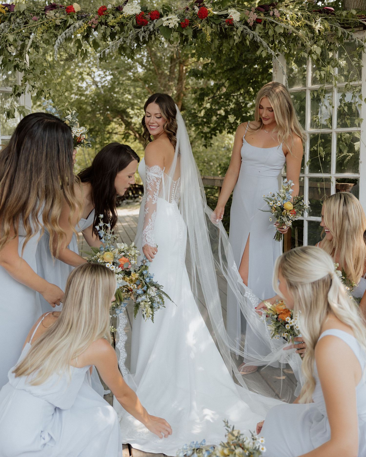 Bridesmaids in light blue dresses gather around bride in white gown with flowing veil during outdoor wedding celebration.