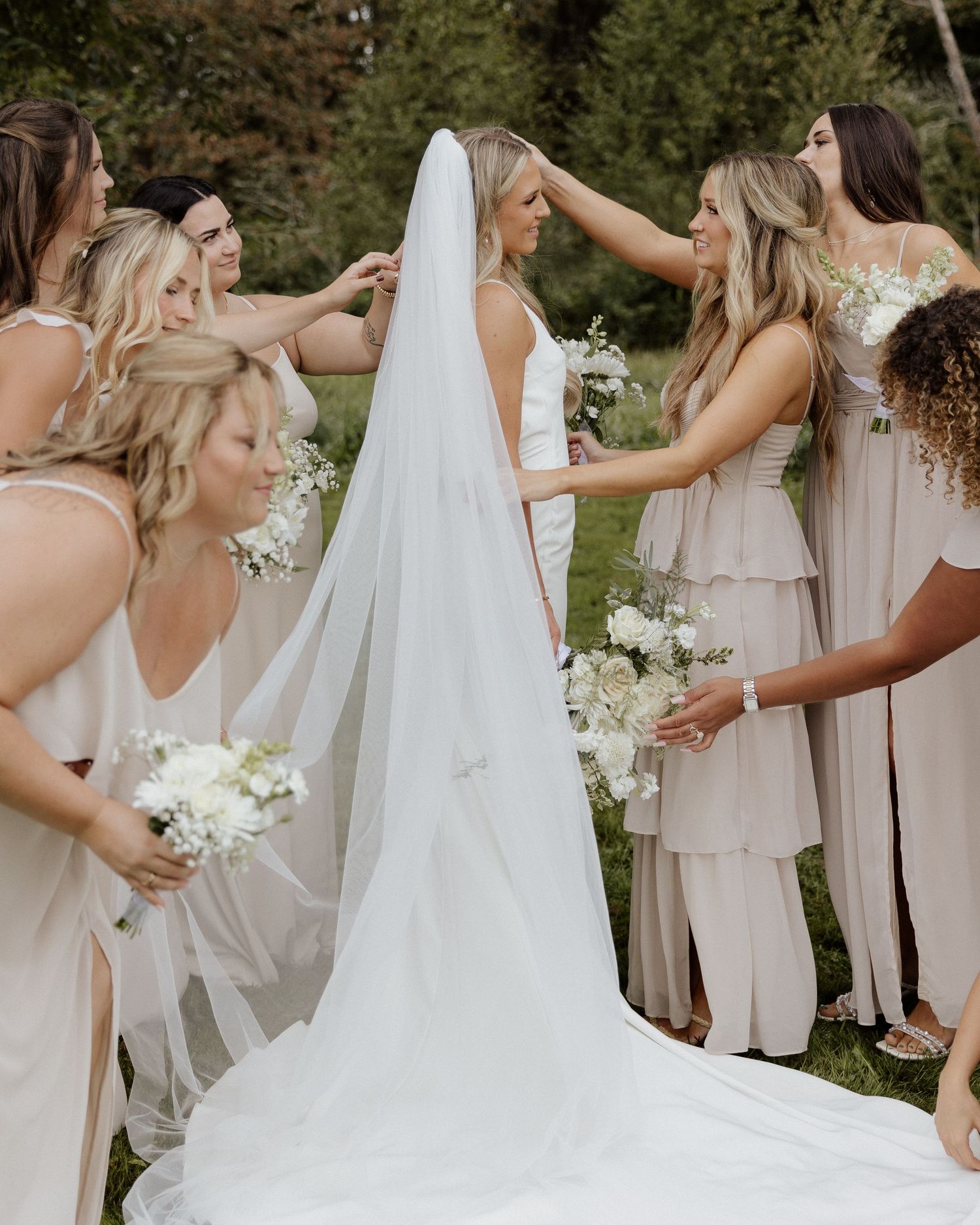 Bridesmaids in blush dresses help adjust the bride's flowing veil during an outdoor photo session.