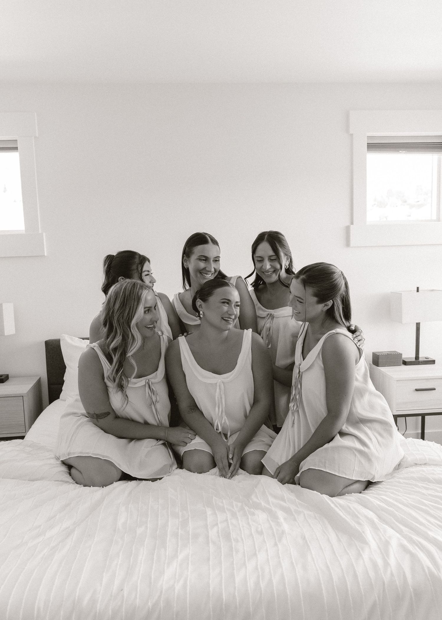 Bridesmaids in white pajamas huddle together laughing on a bed during bridal preparations.
