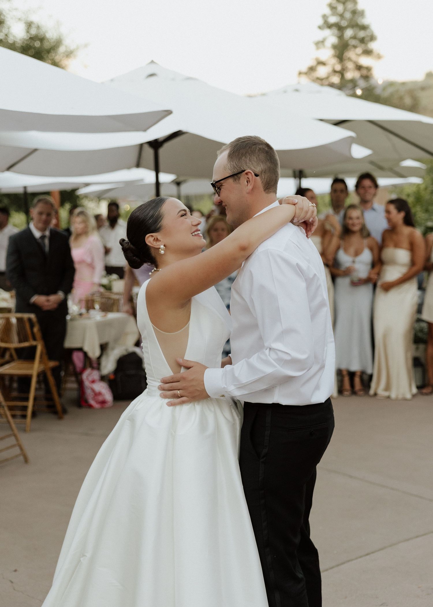 Smiling bride and groom share first dance outdoors at wedding reception with guests watching.