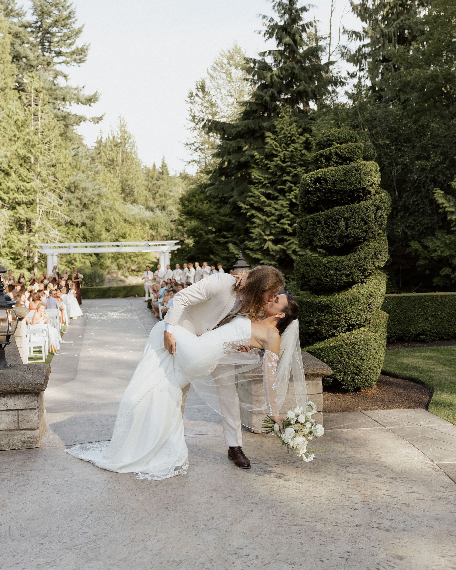 Groom dipping bride for romantic kiss along outdoor wedding ceremony aisle lined with guests.