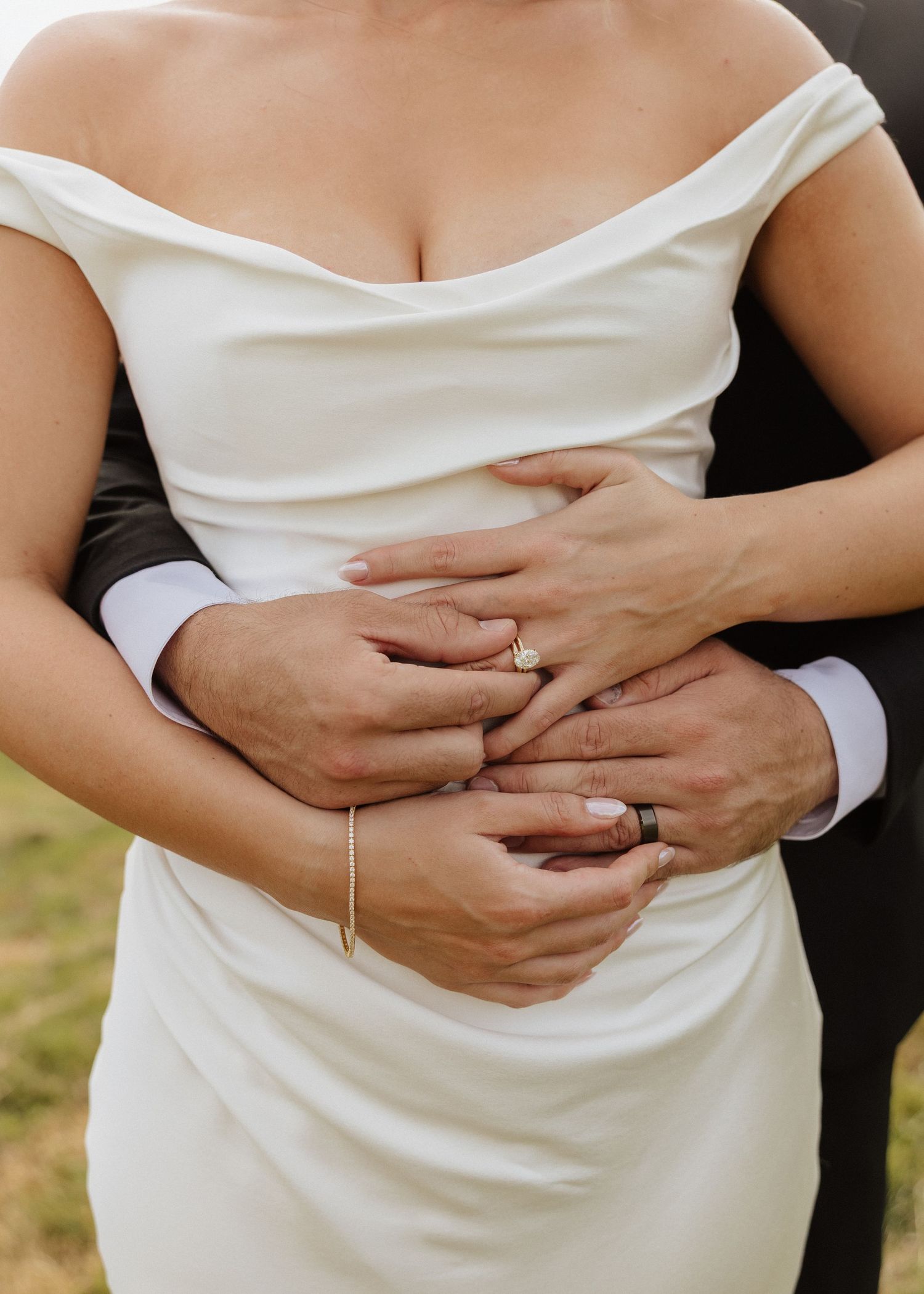 Close-up of bride and groom's hands showing wedding rings, groom embracing bride in white satin gown.